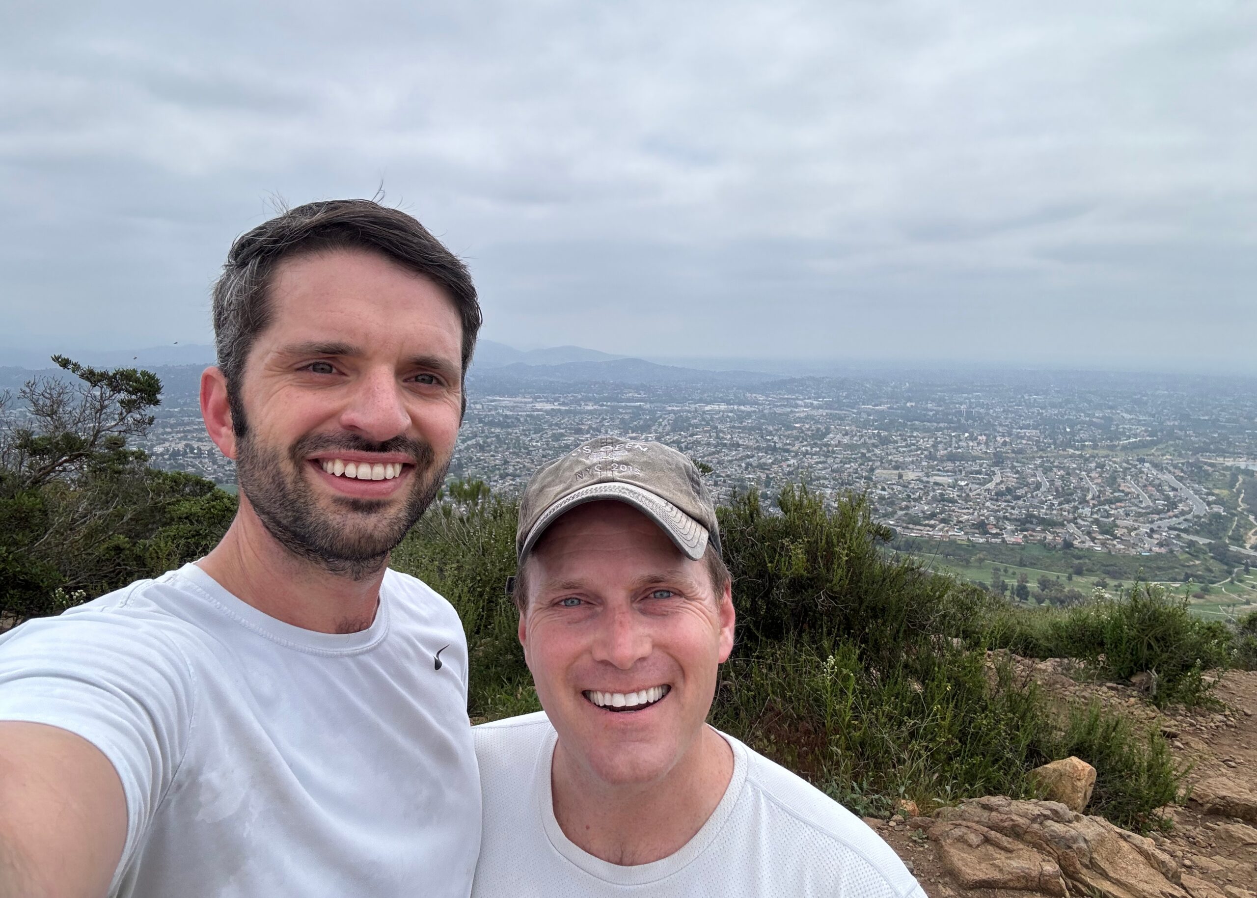 Two men smiling for a hillside selfie with a valley and city spread out below them under a cloudy sky in the background, rocky terrain in foreground.