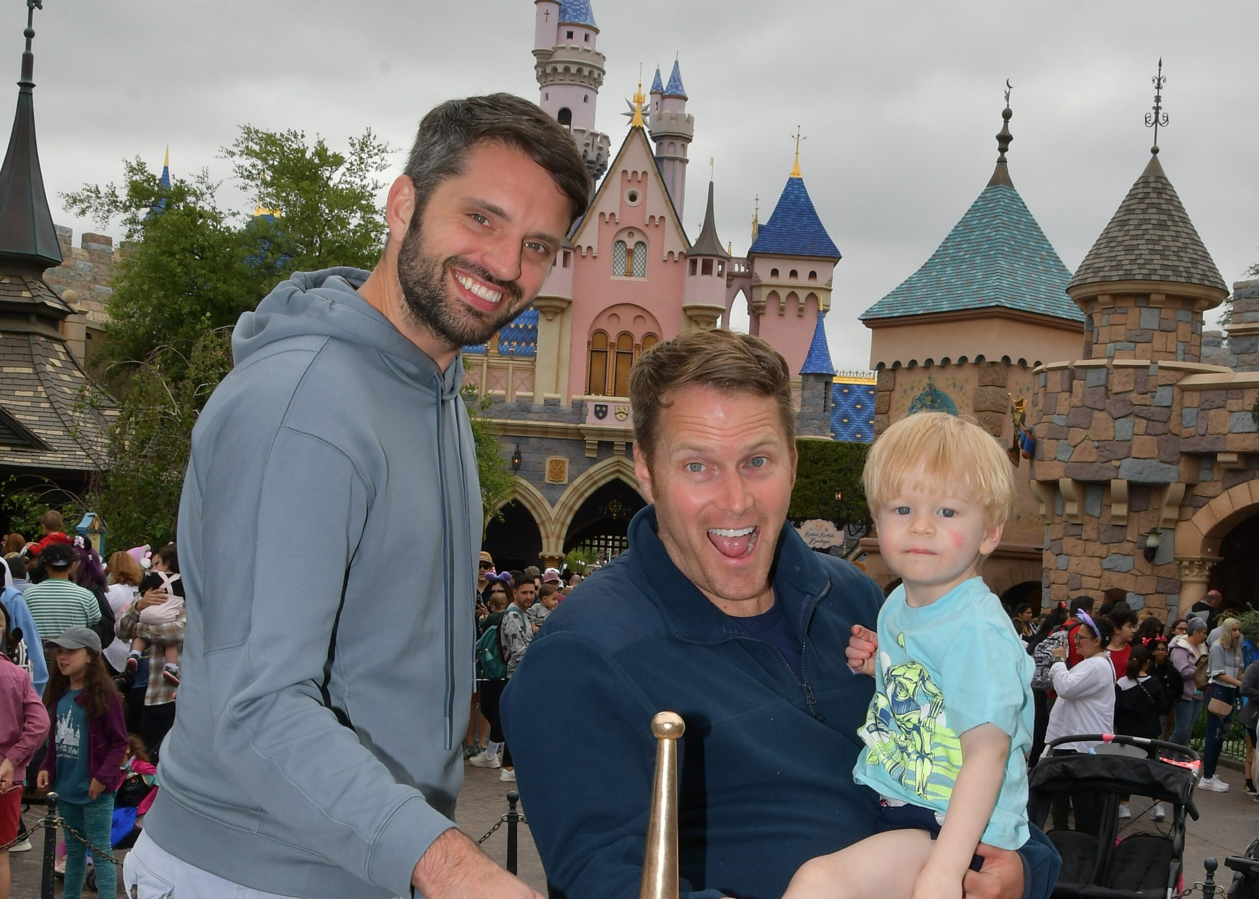 Man in gray hoodie and a man with a child pose for a photo in front of a pink castle at an amusement park.