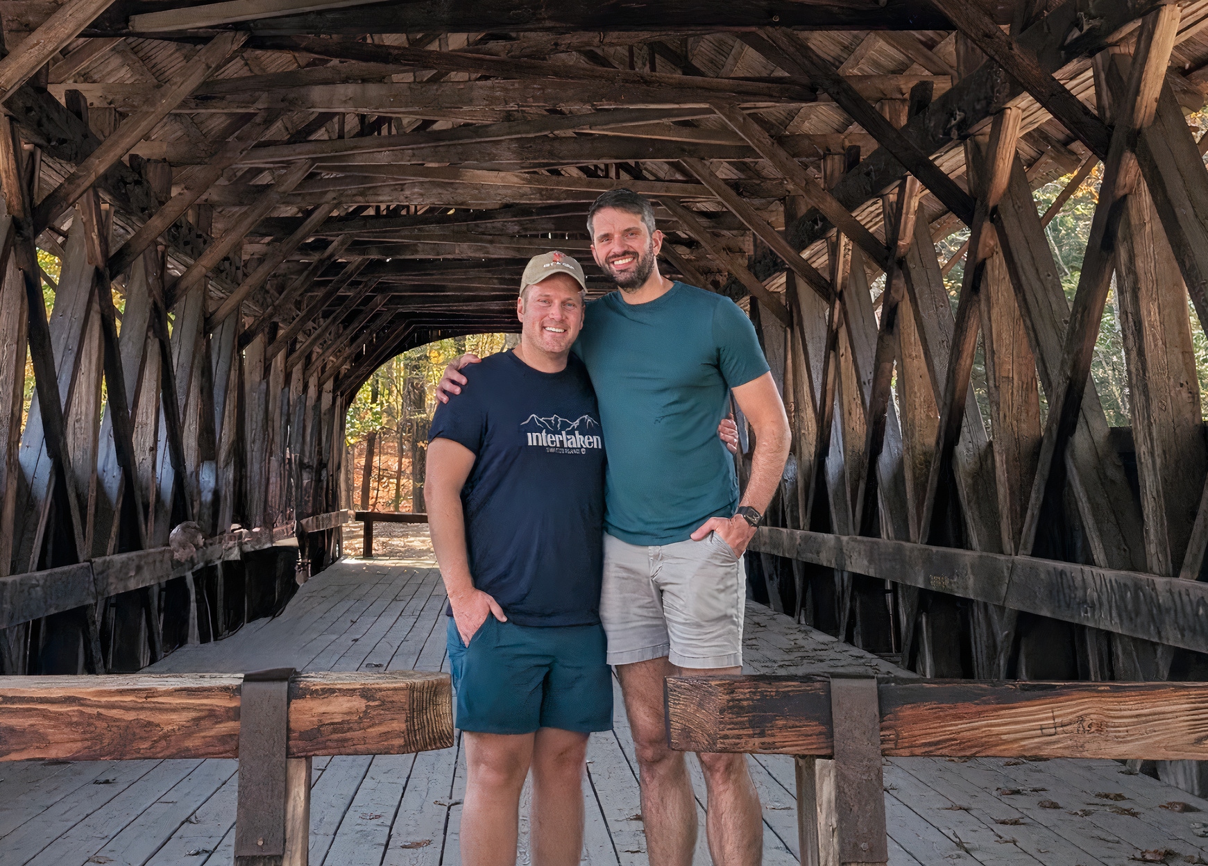 Two men stand arm-in-arm inside a rustic wooden covered bridge, smiling at the camera.