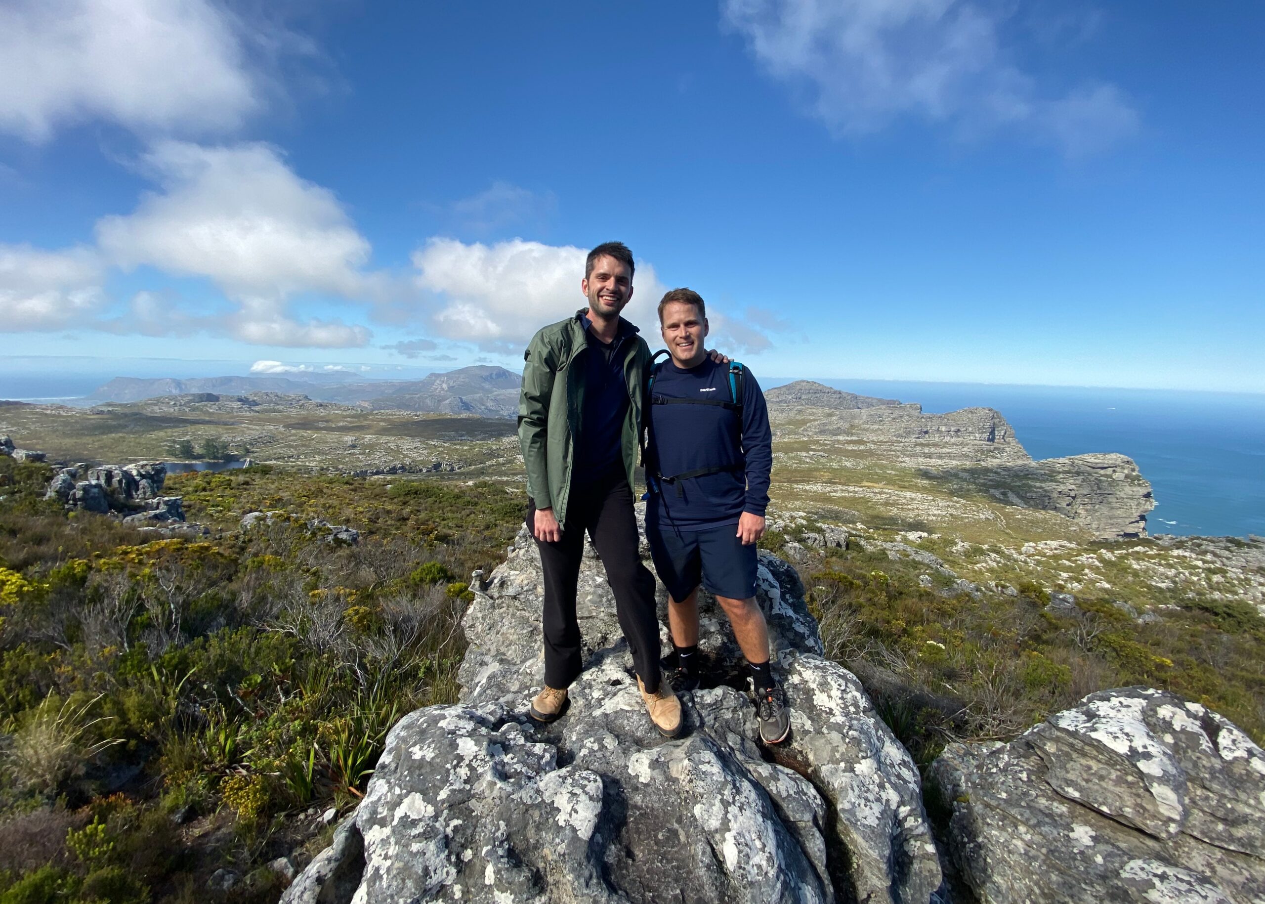 Two men in outdoor gear posing together on jagged rocks at a coastal overlook, with blue sky and distant sea in the background.