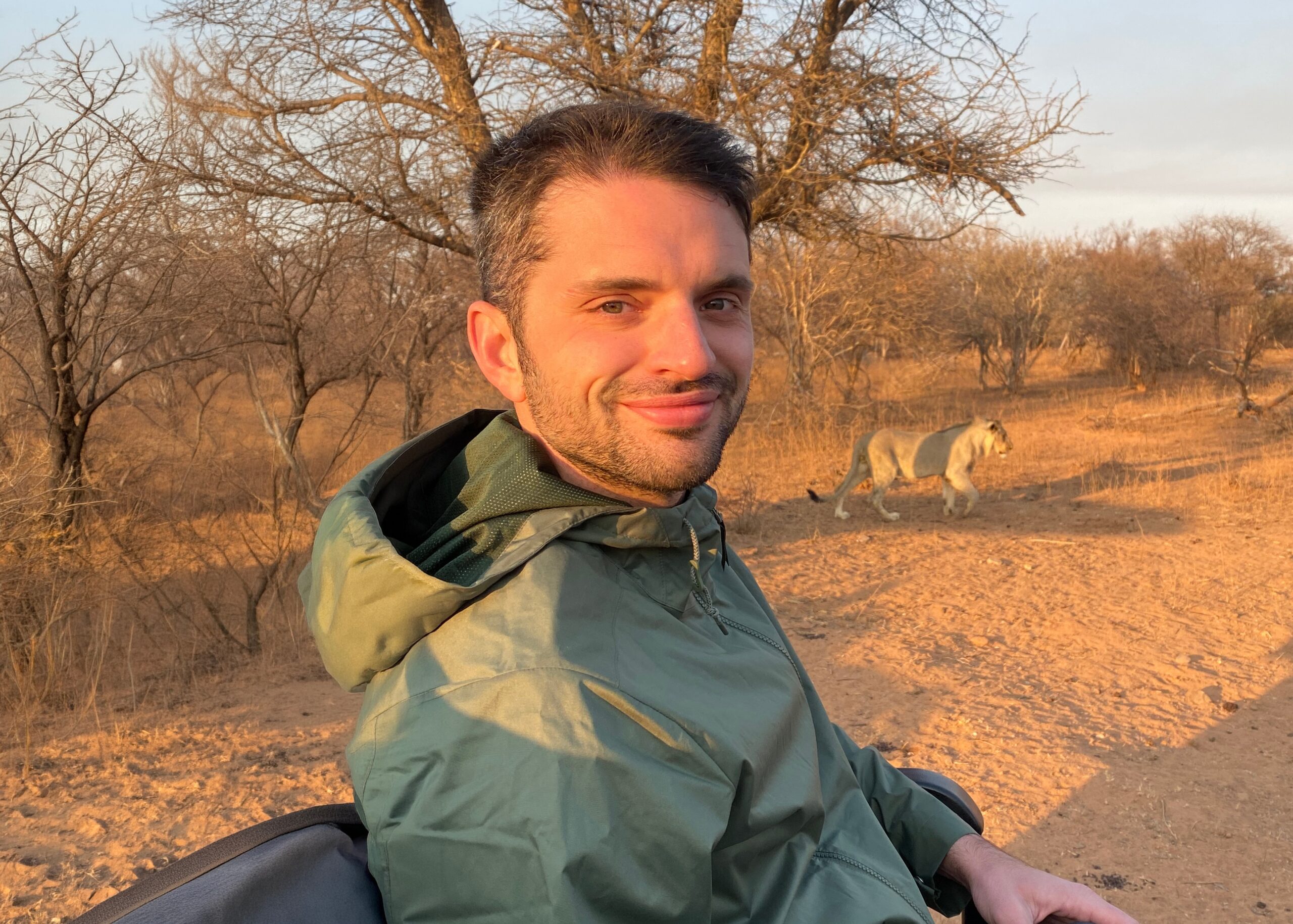 Selfie of a man in a green jacket on a dry savannah, with a lion walking in the background.