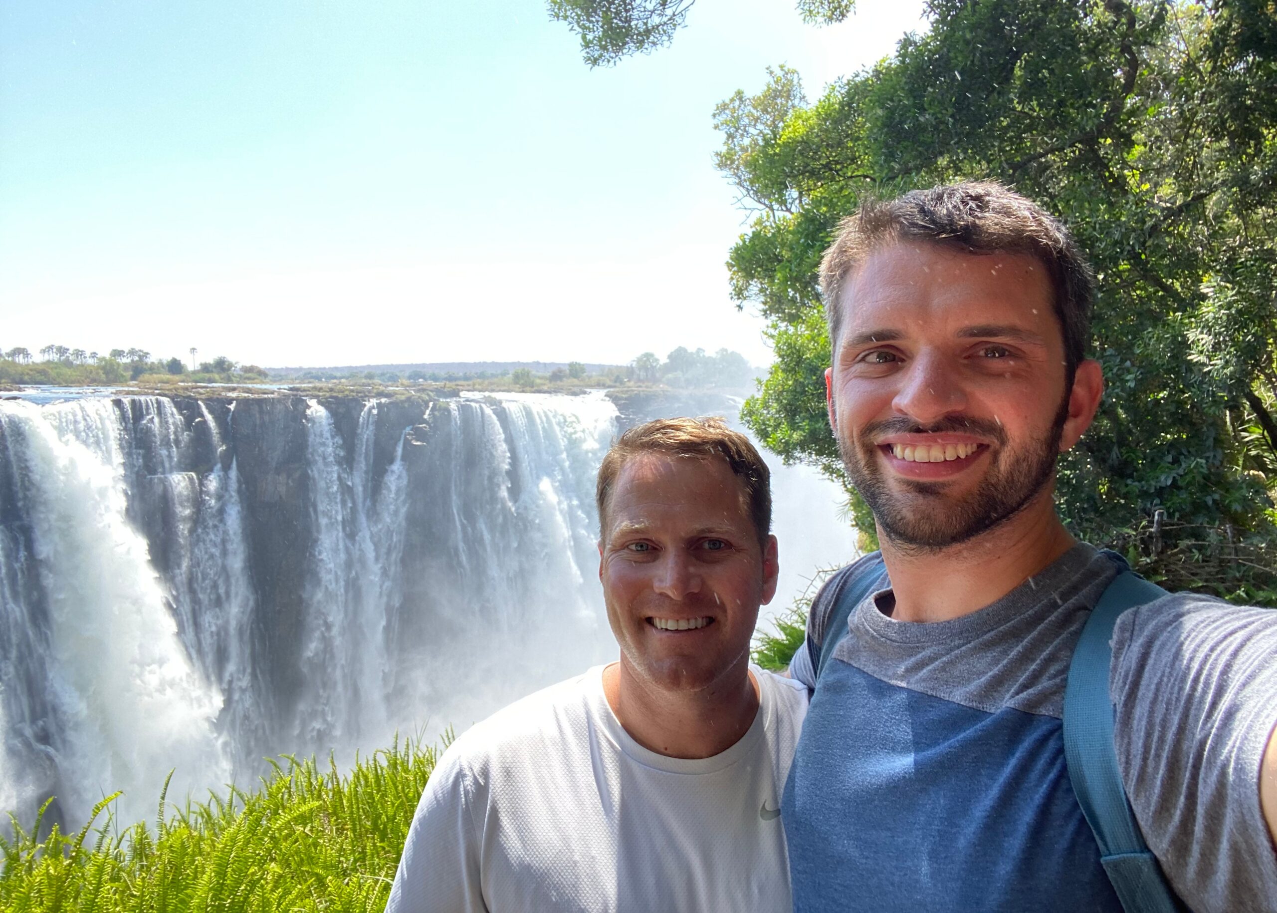 Two men taking a selfie in front of a wide waterfall with mist and lush green plants nearby.