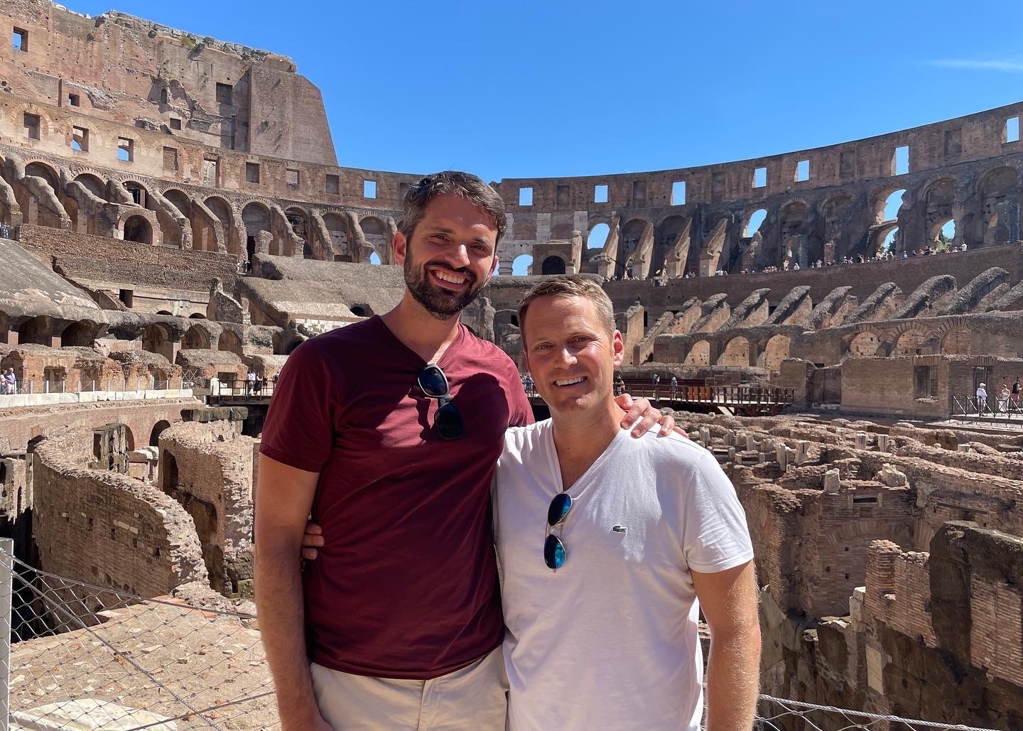 Two men smiling and posing inside the ancient Roman Colosseum with arches and stone seats behind them, wearing casual t‑shirts and sunglasses.