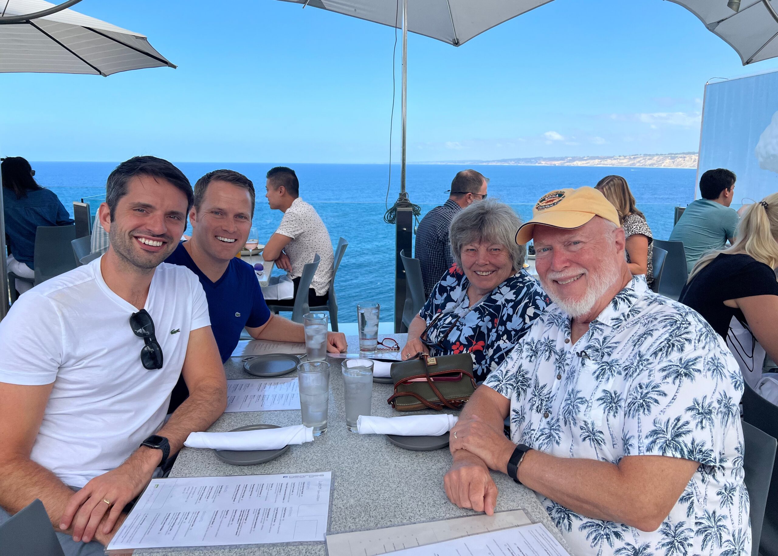 Smiling group of diners at an outdoor seaside restaurant with the ocean and coastline in the background.