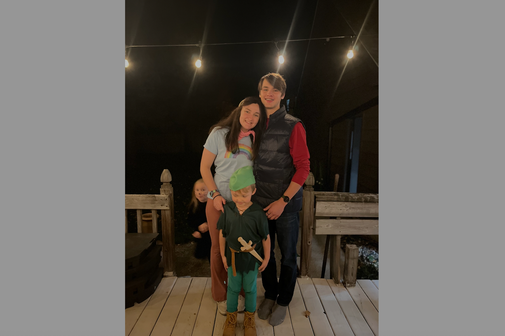 A family stands together on a wooden deck at night under string lights, smiling at the camera.