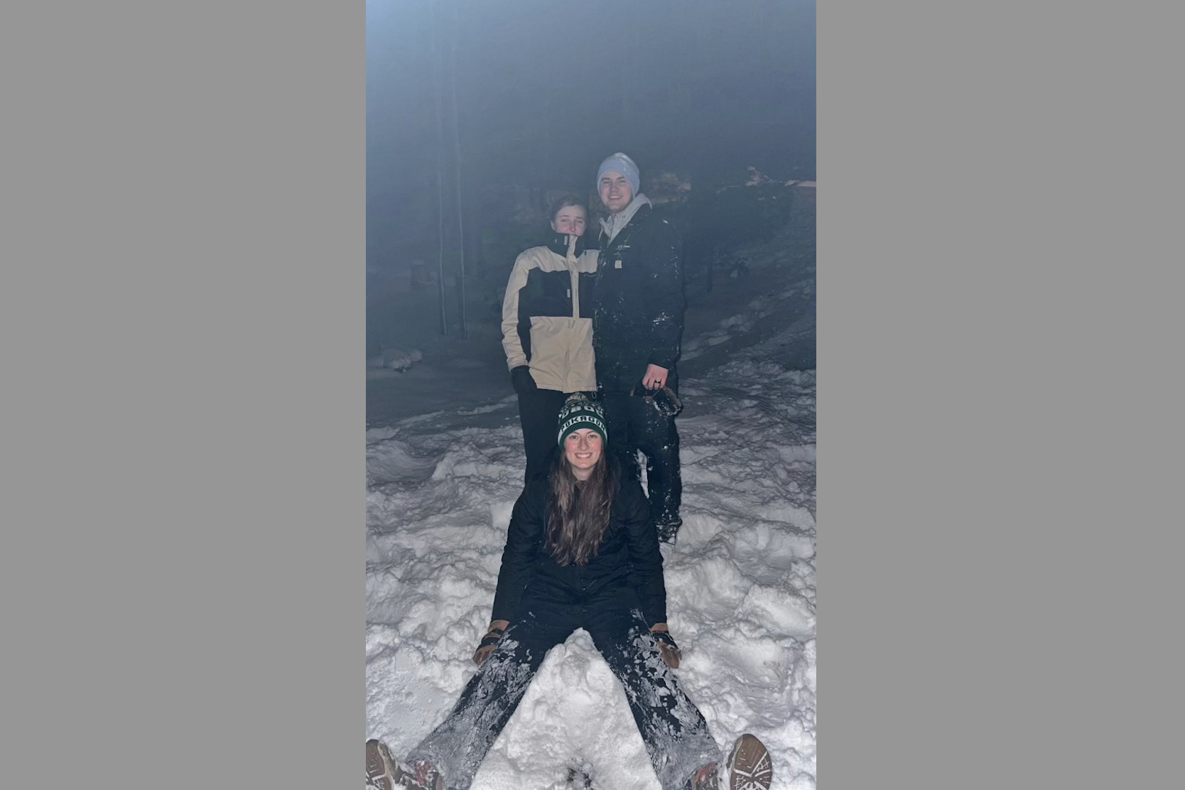 Three friends posing in a snowy night scene, two standing and one kneeling in front, all dressed in winter coats and hats.