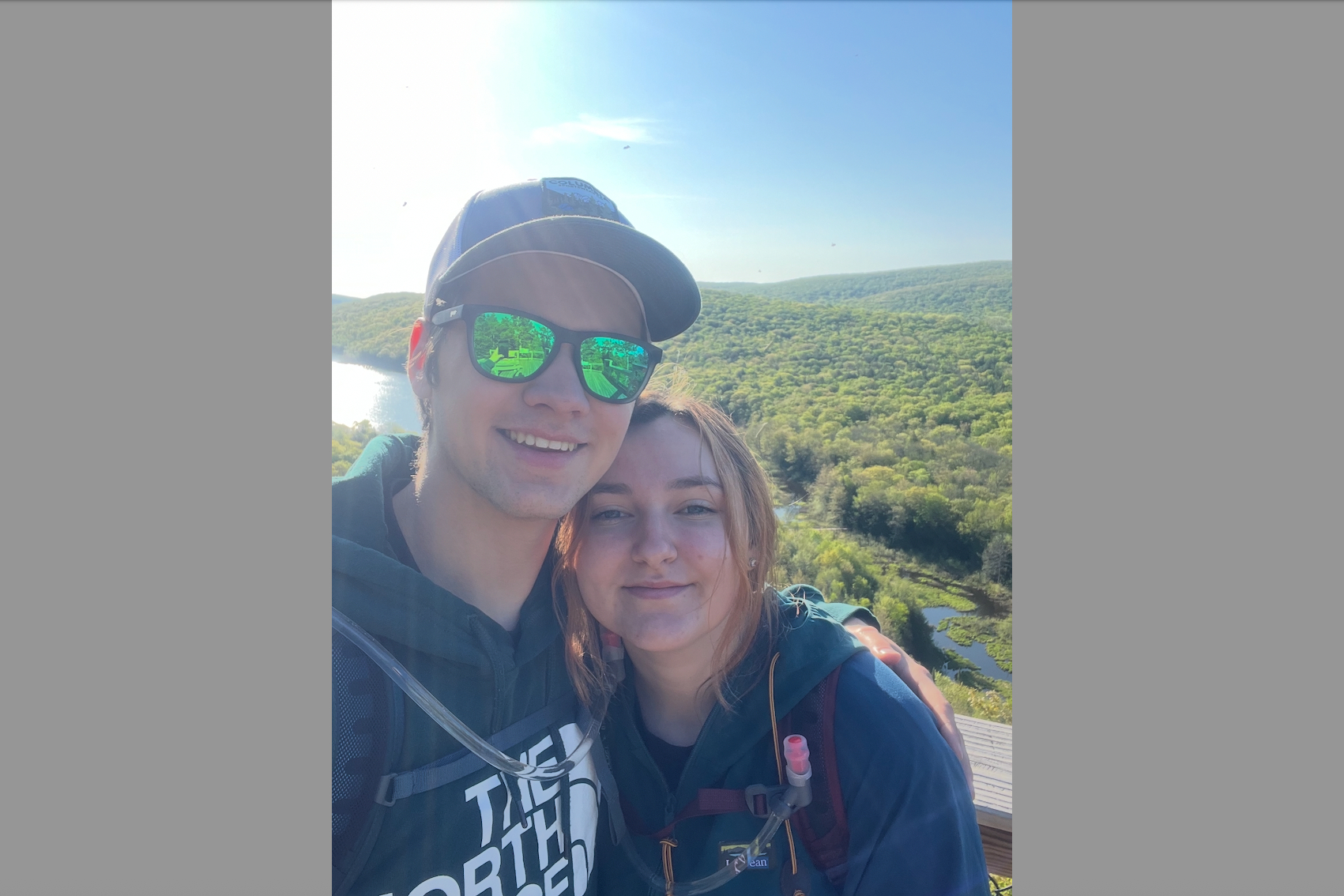 Two friends pose for a selfie on a sunny overlook with a forested valley and river in the background.