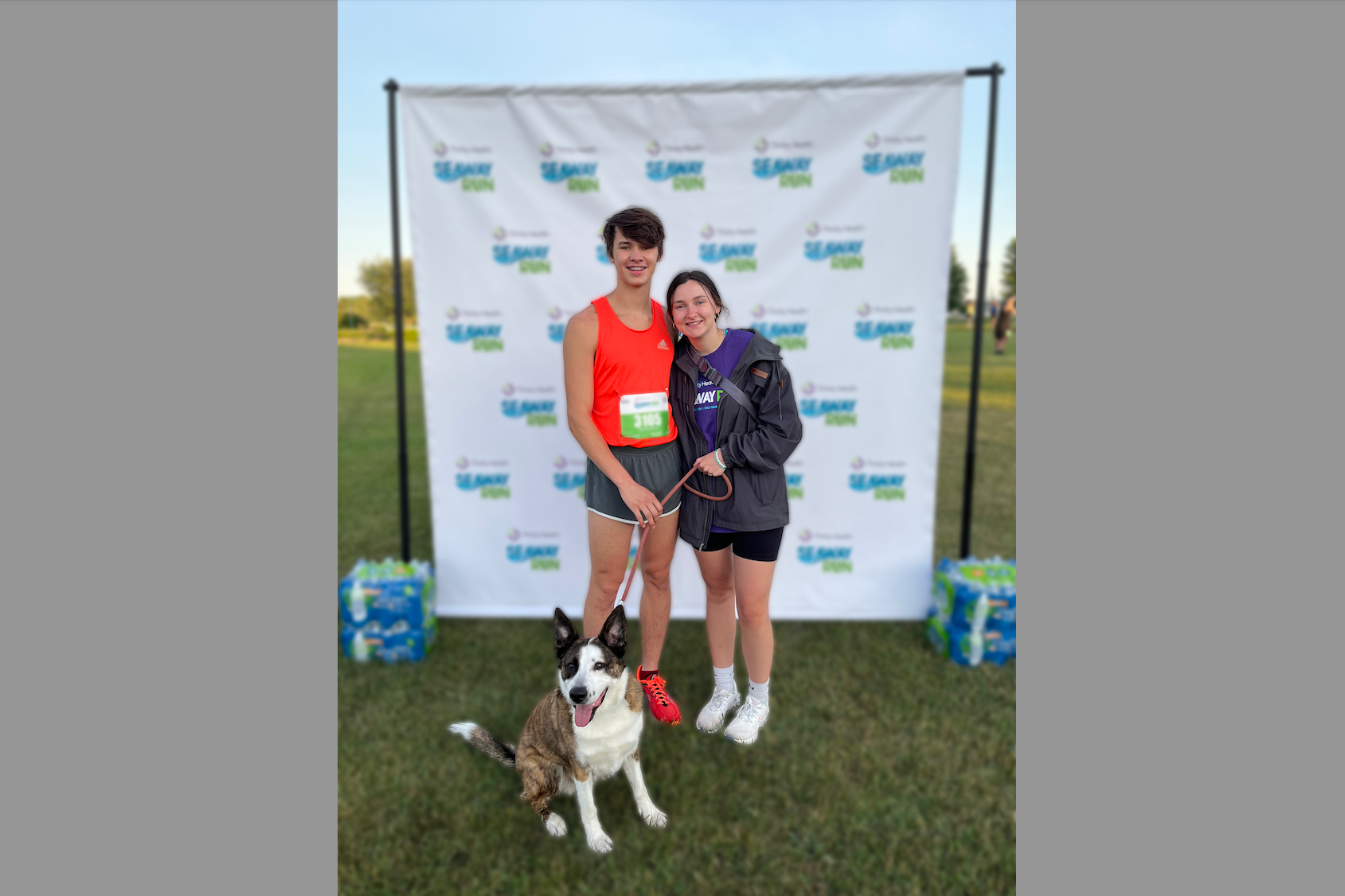 Two young runners pose with their leashed dog in front of an outdoor race backdrop and banners, smiling at the camera with race bibs visible on their outfits.