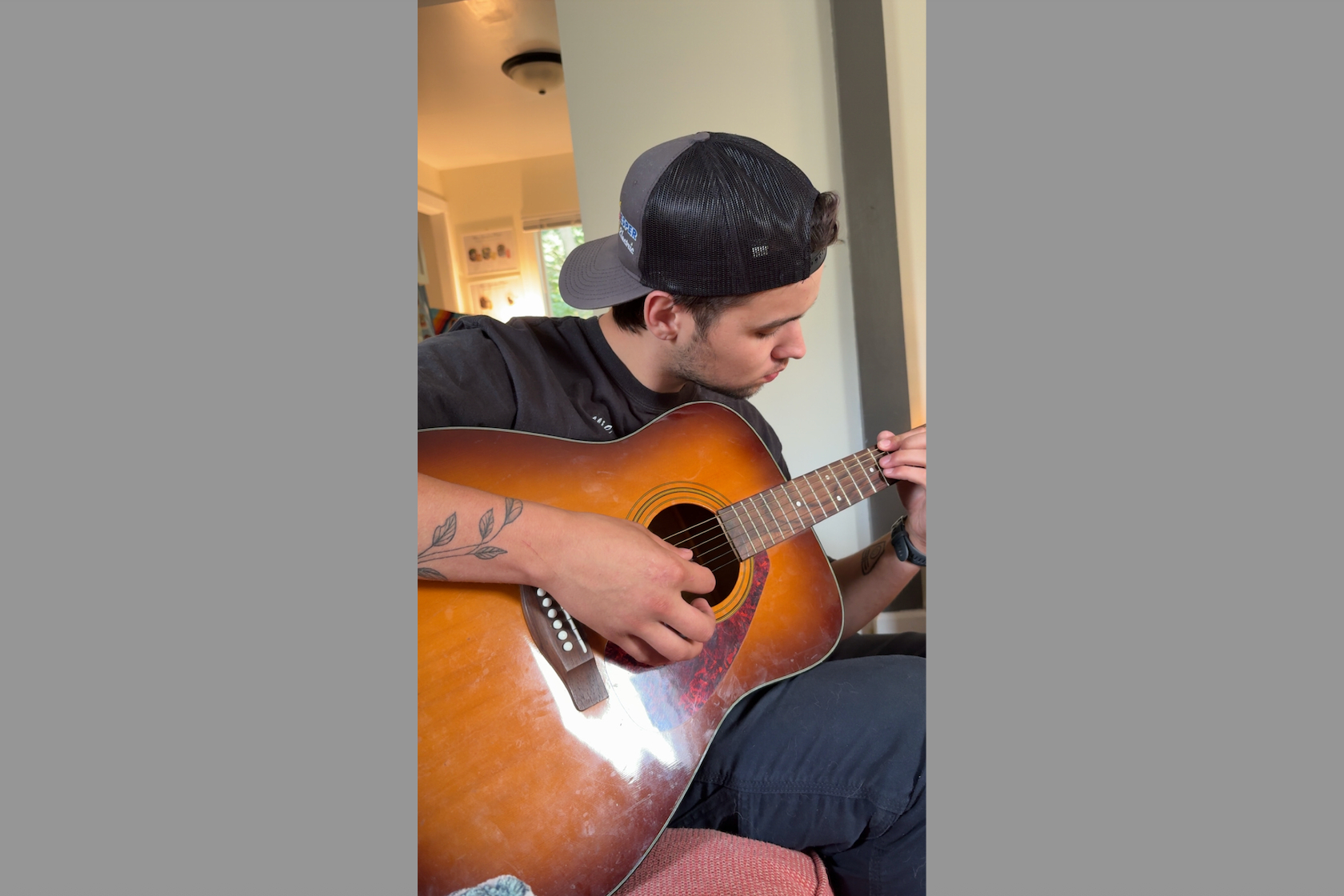 Young man wearing a baseball cap plays an acoustic guitar indoors, focusing on strumming the strings.