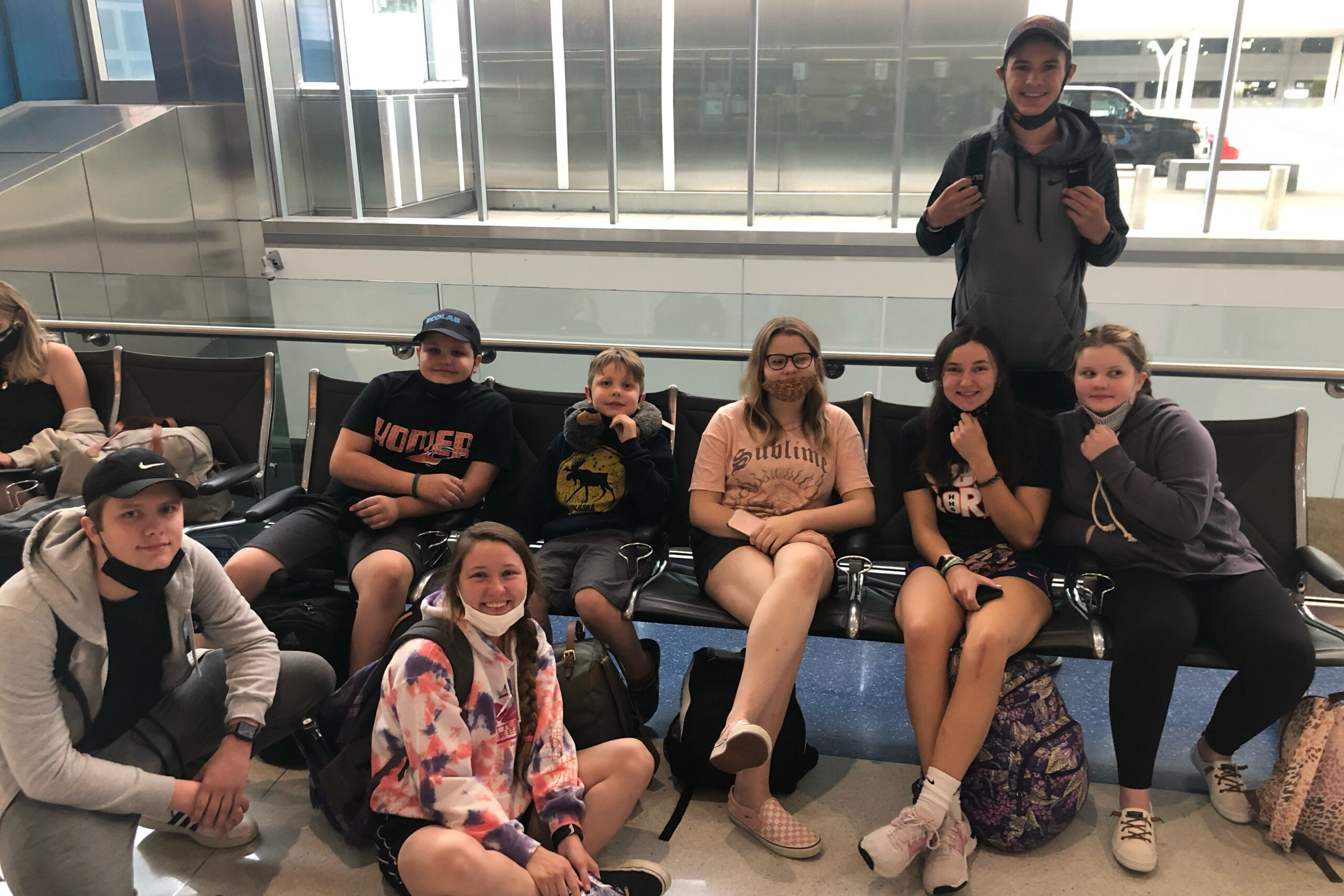 Group of smiling teens and preteens seated in a row of chairs at an airport, one standing with backpack straps visible behind them