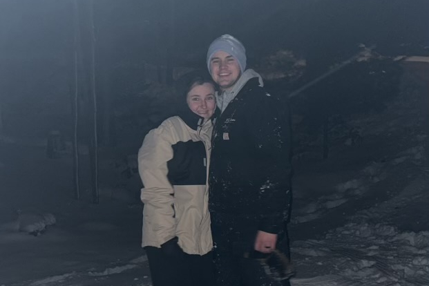 Smiling couple in winter coats standing close in the snowy night, arms around each other.