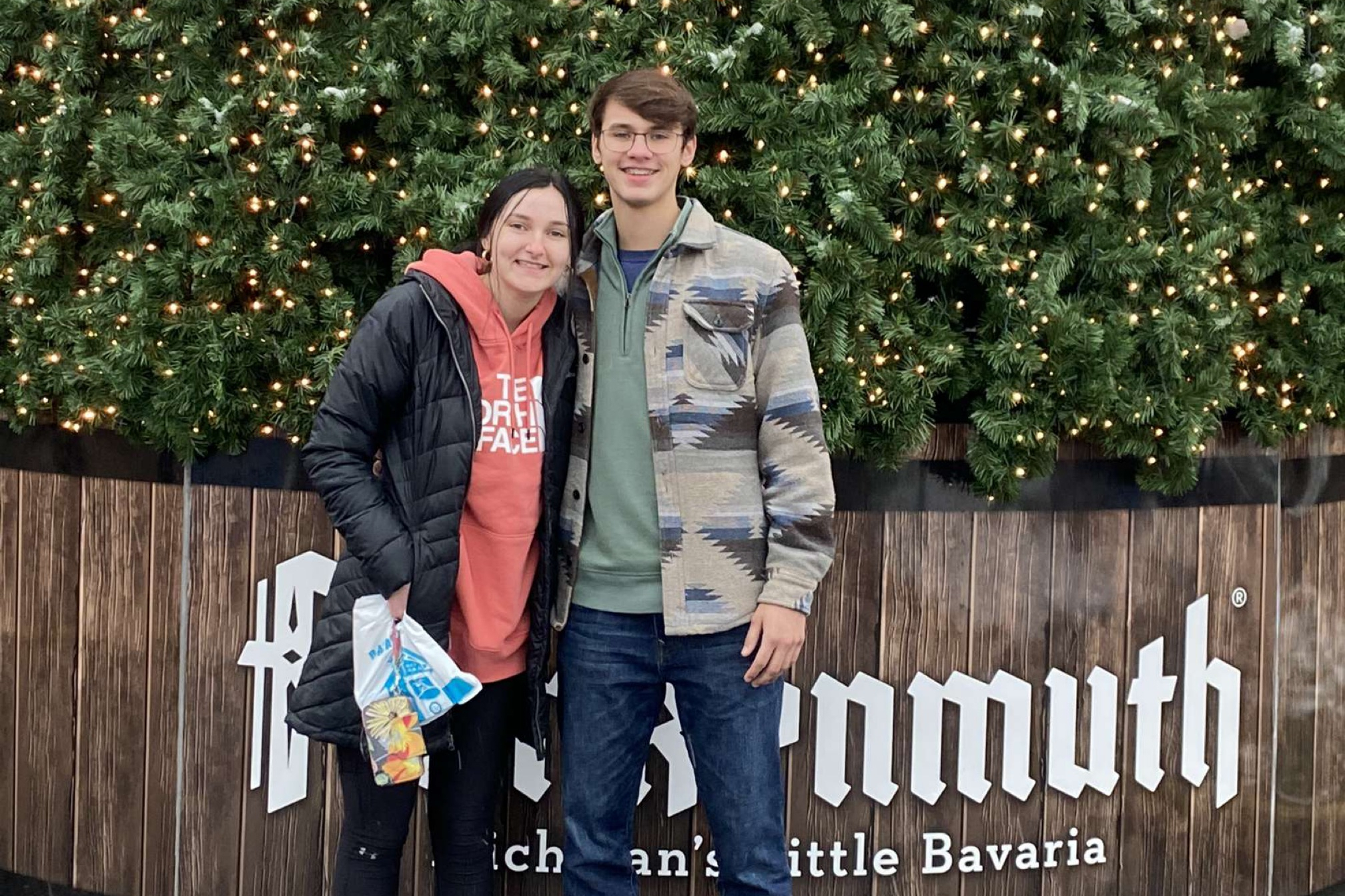 Two friends pose together in front of a lit evergreen backdrop with string lights and a wooden fence behind them, wearing jackets.