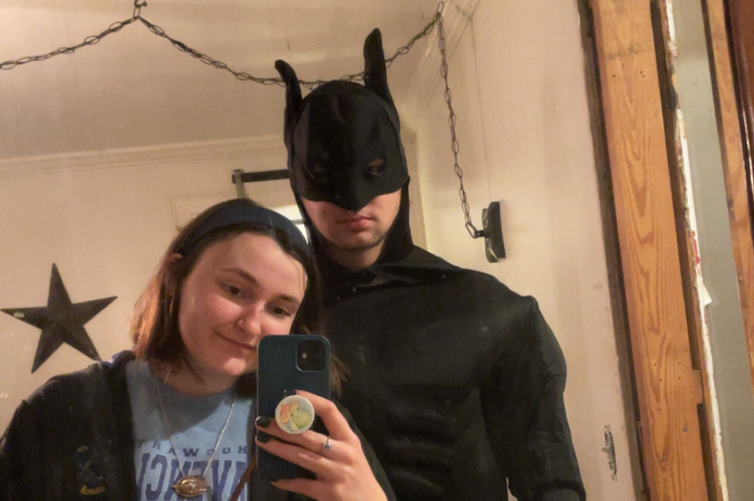 Young woman takes a mirror selfie with a man dressed as Batman—mask and black suit—standing behind her in a room with a star decoration on the wall.