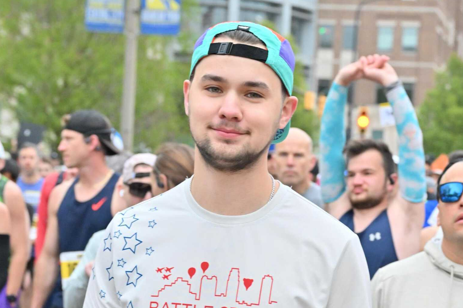 Smiling young man in a turquoise and purple backward-cap at a street race, with other runners behind him in an urban setting.