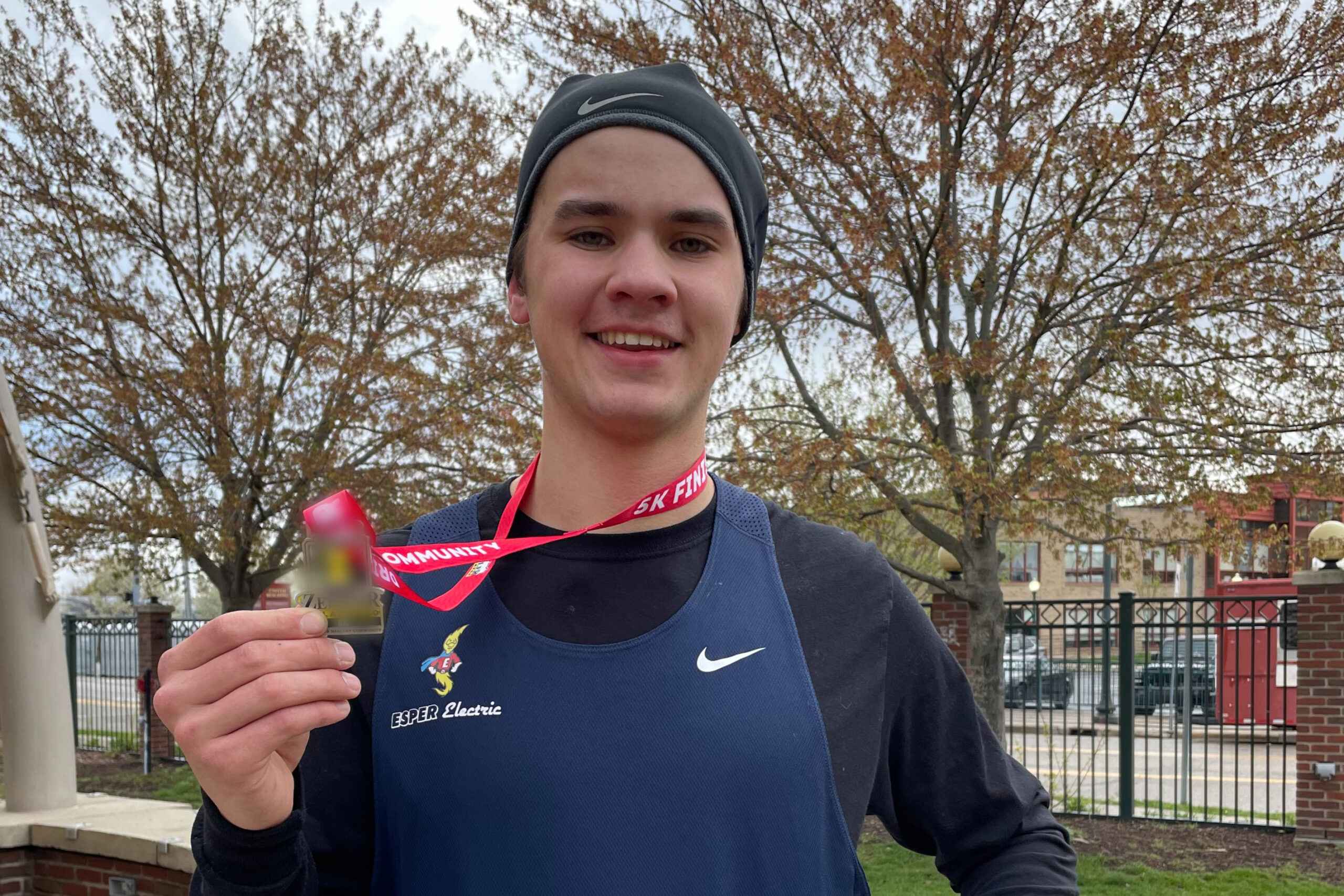 Smiling runner wearing a black beanie and blue athletic singlet, holding up a medal with a red ribbon outdoors with trees in the background.