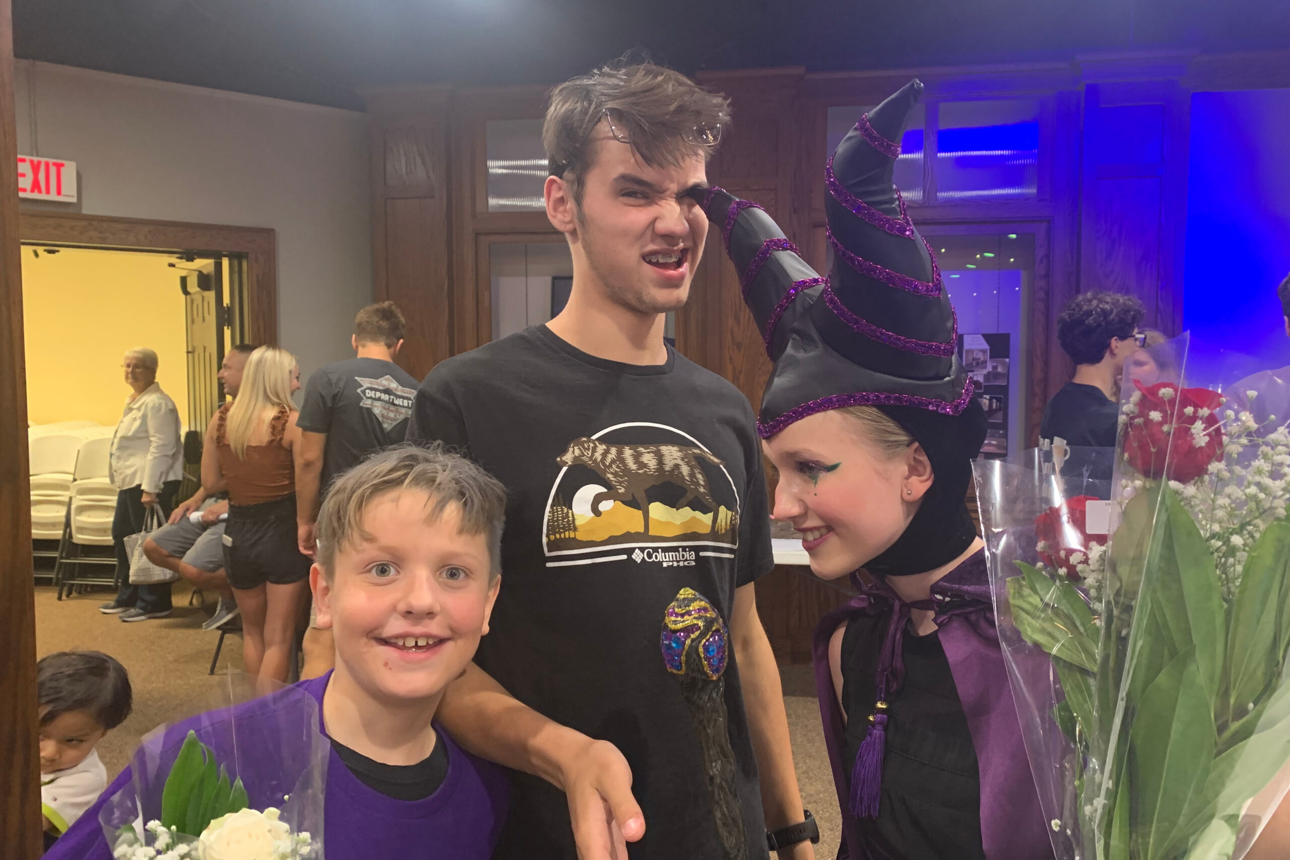 Joyful group photo at an indoor event: smiling kids with a person in a purple-horned costume and others in the background near flowers and exit sign.