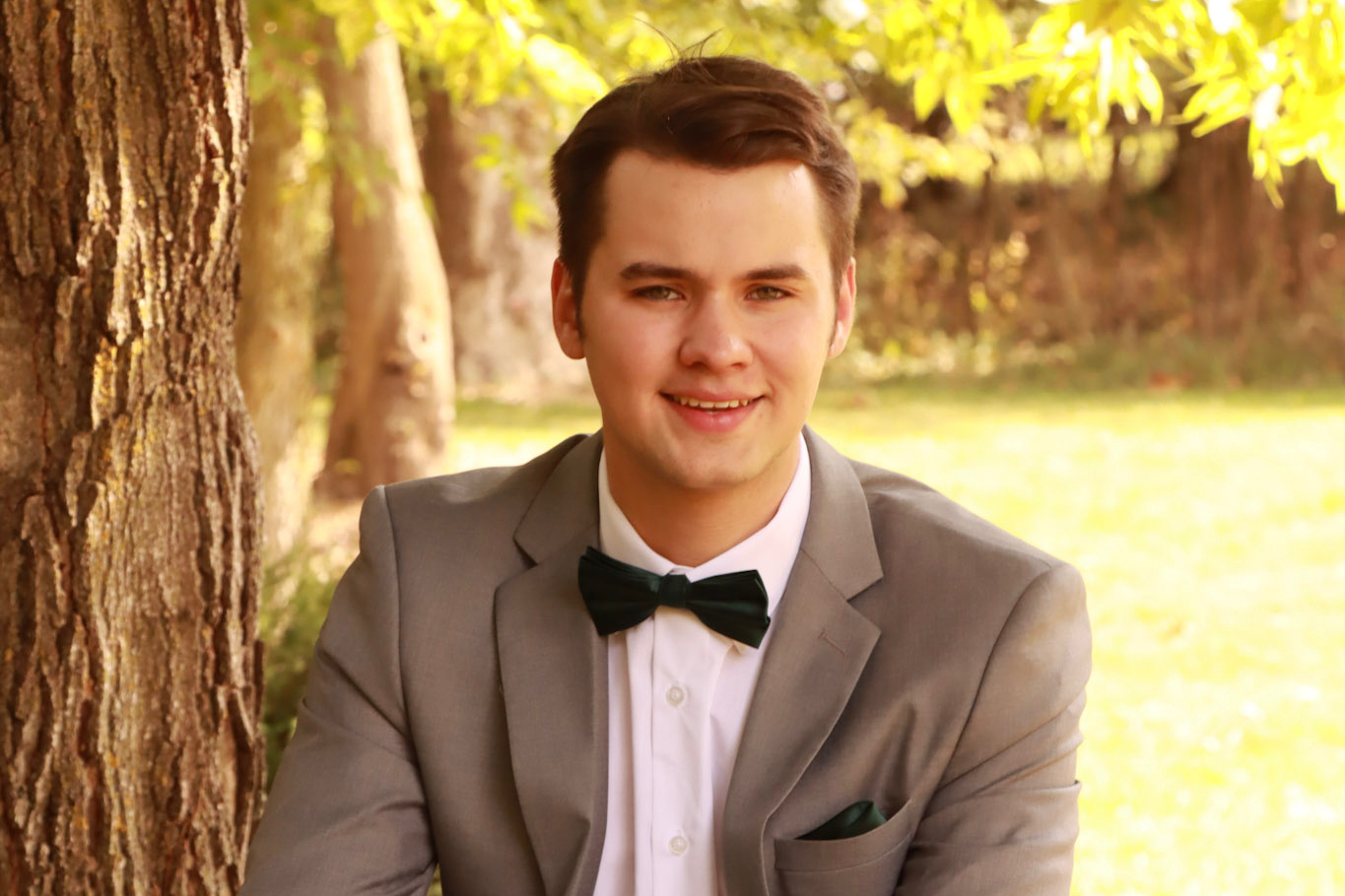 Young man in a gray suit and black bow tie smiles while leaning against a tree in a sunlit park.
