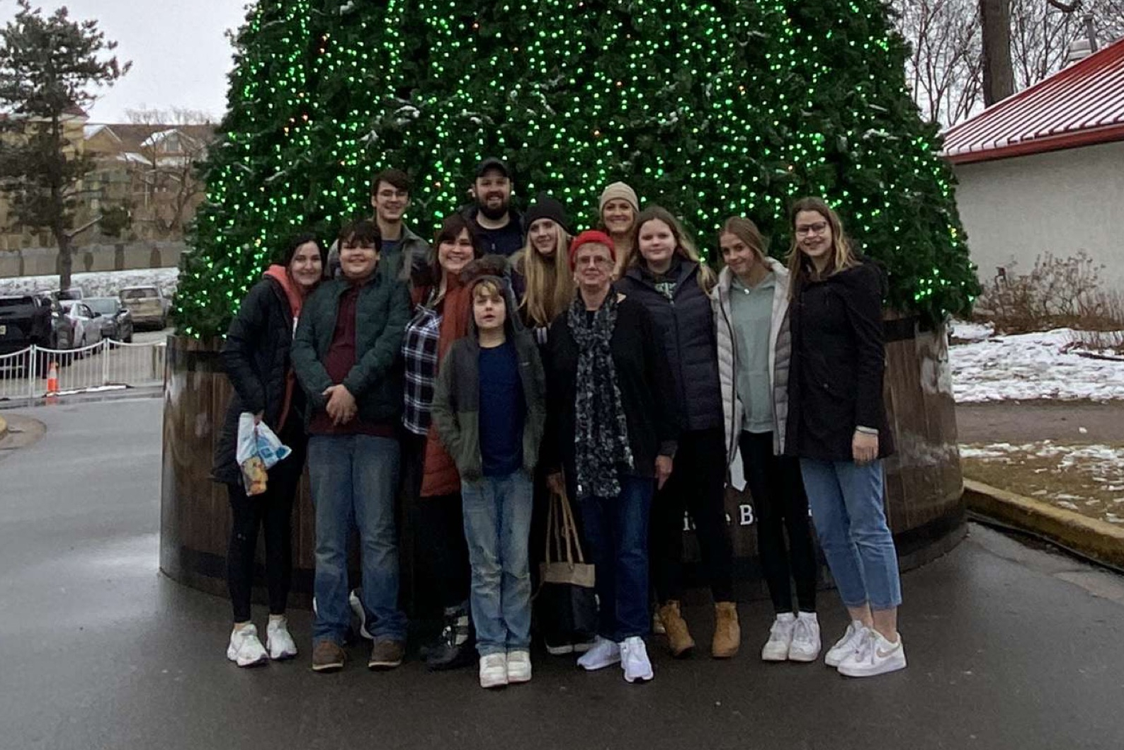 Group of people posing in front of a large, festively lit Christmas tree outdoors in winter.