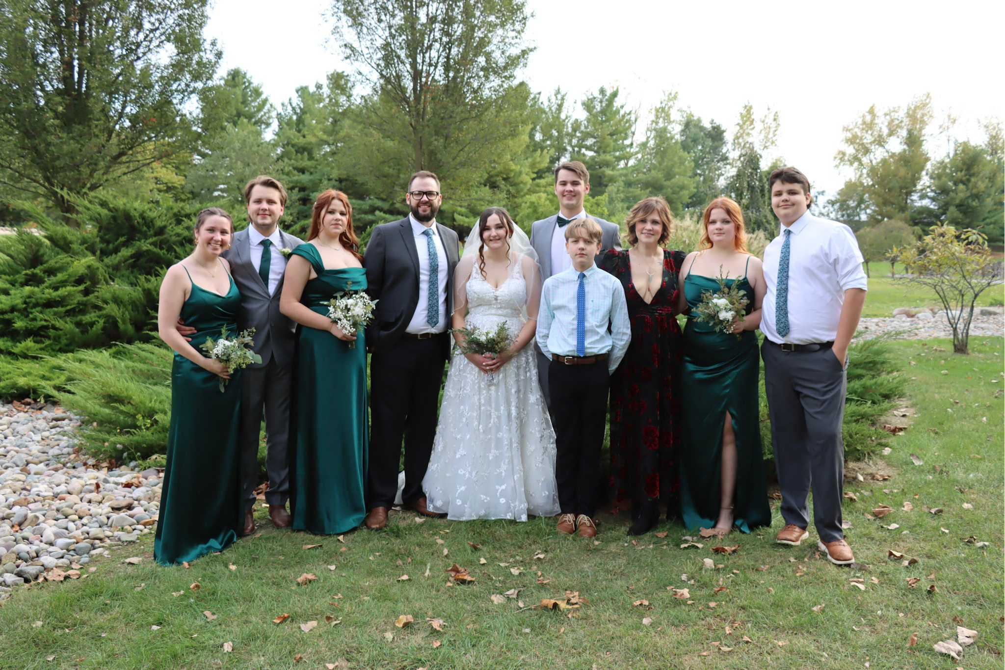 Outdoor wedding party posing together on a lawn with trees; bridesmaids in green dresses, groomsmen in suits, the bride in a white lace gown, and a young boy in a blue shirt.