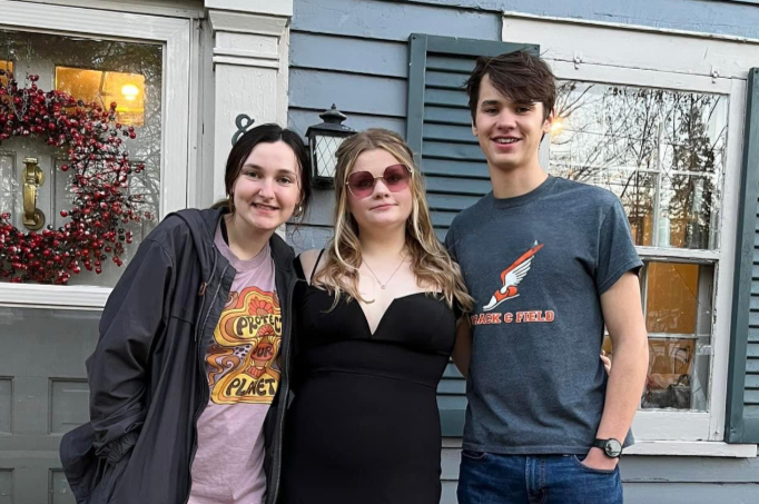 Three friends posing together outside a blue house, smiling at the camera, one in a black dress, one in a bomber jacket, the other in a t‑shirt and jeans with sunglasses on head accessories nearby.