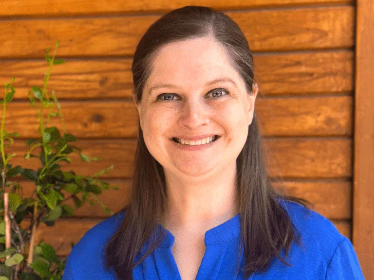 Smiling woman with long dark hair in a blue blouse, standing in front of a wooden panel wall with a plant to her left.