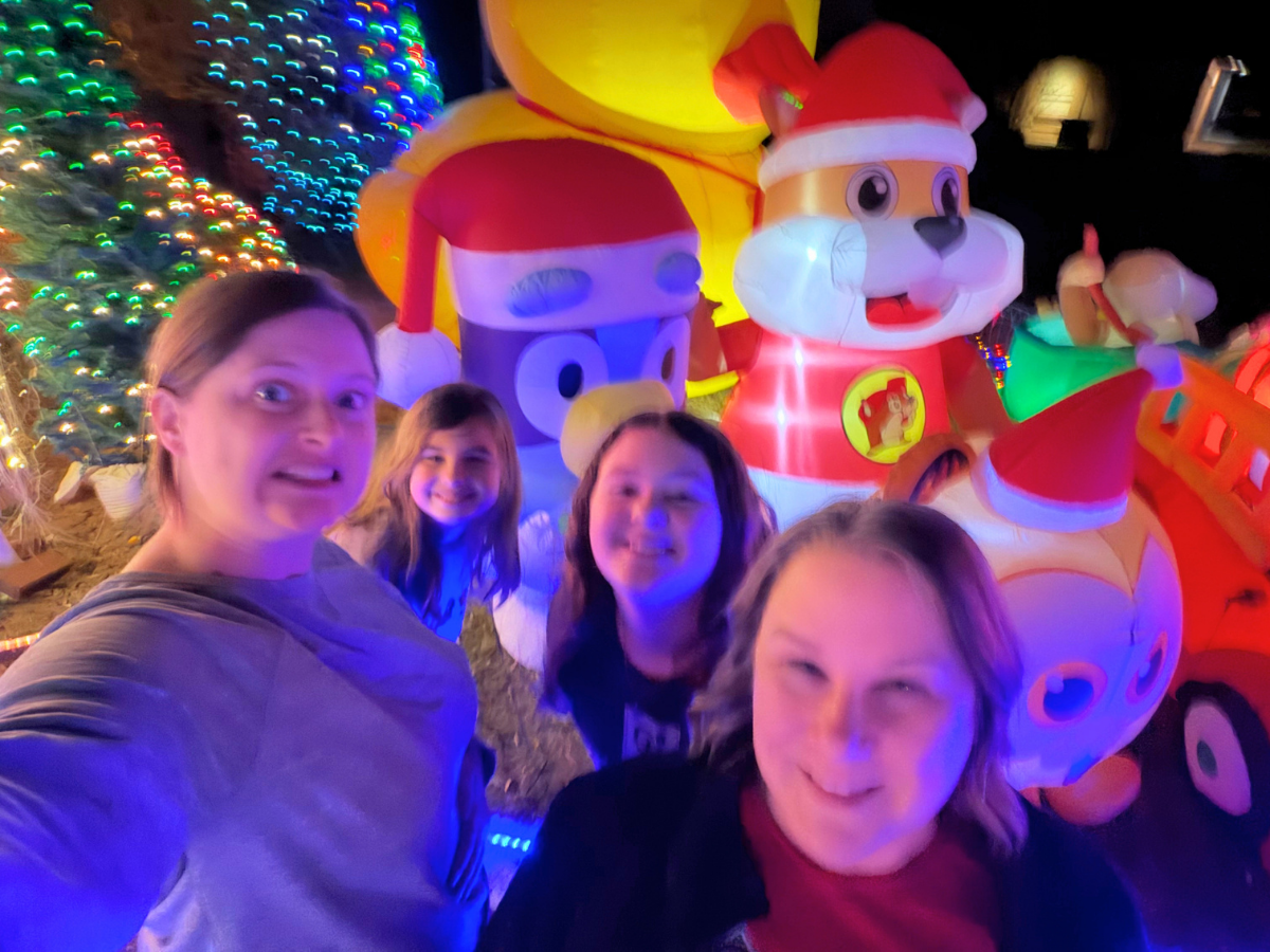 Four friends take a selfie in front of a colorful inflatable Christmas display with Santa-hatted animals at night.