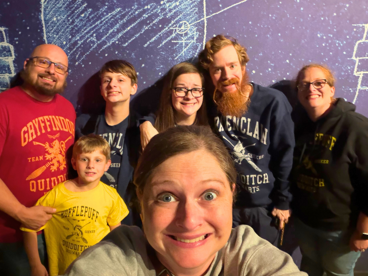 Seven people pose for a group selfie in front of a purple chalkboard wall; the woman in front holds the camera.