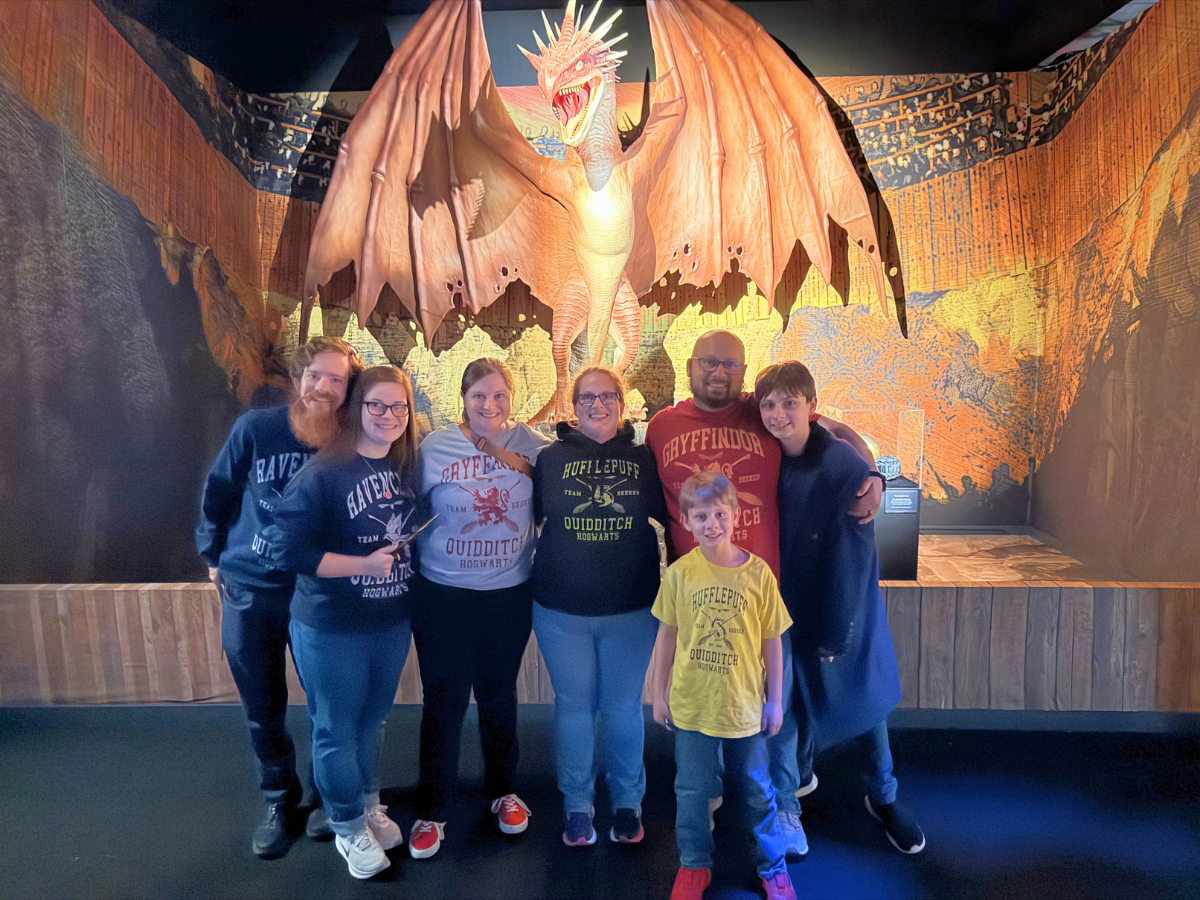 Group of seven people posing together in front of a large dragon sculpture in a museum exhibit, wearing Hogwarts-themed shirts.