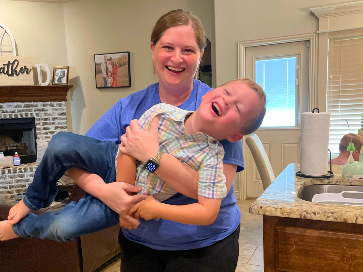 Woman in a blue shirt playfully holds a laughing child while swinging him in a kitchen dining area.