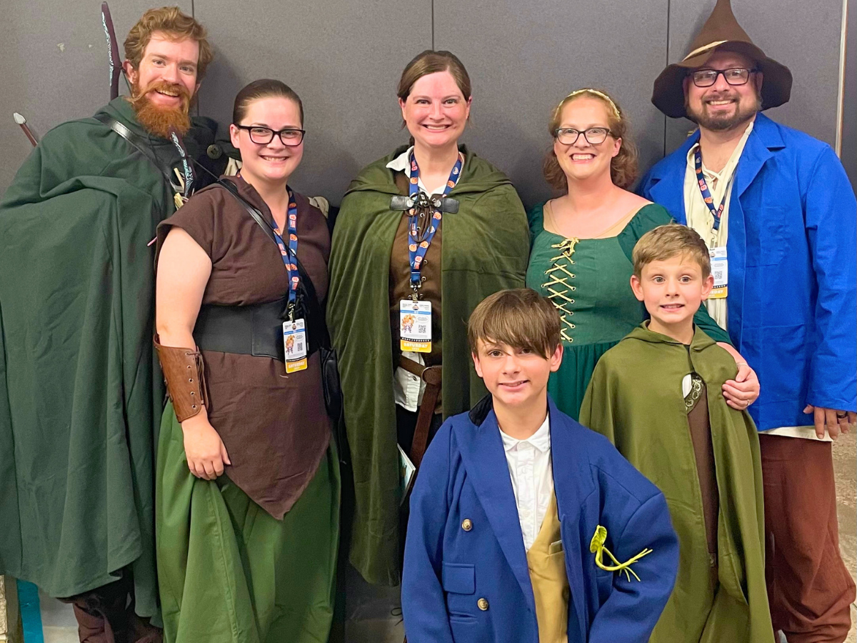 Group of eight people in medieval/fantasy costumes posing together for a photo, wearing cloaks, tunics, and a wizard hat, with lanyards and badges visible.