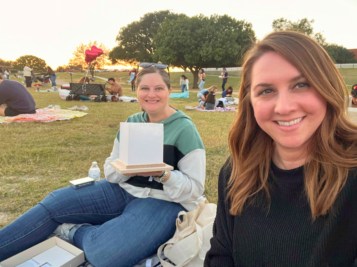 Two women sit on a grassy park at sunset; one holds a small white box on a wooden base while others picnic in the background.