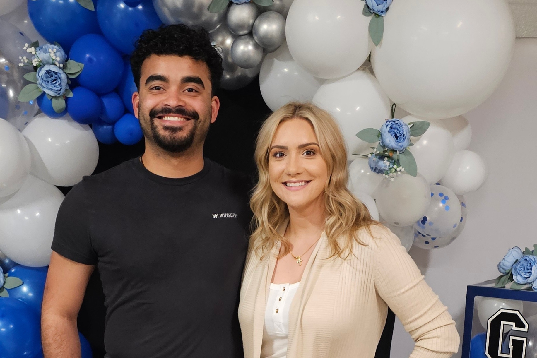 Smiling man in a black t-shirt and smiling woman in a beige cardigan pose in front of a blue-and-white balloon arch with blue roses and greenery."