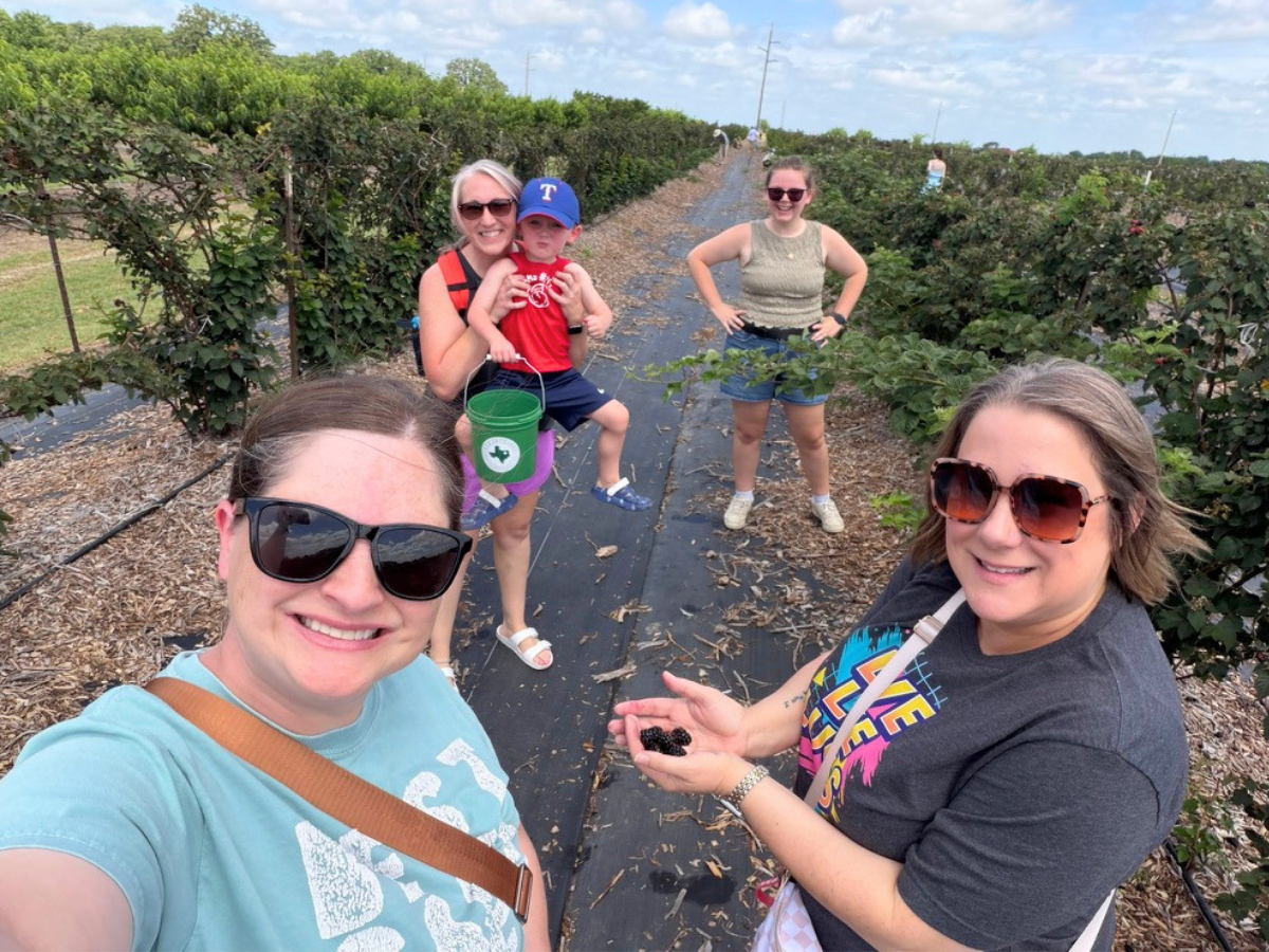 Group of adults and a child picking blueberries on a farm row, smiling for a selfie with buckets of berries nearby