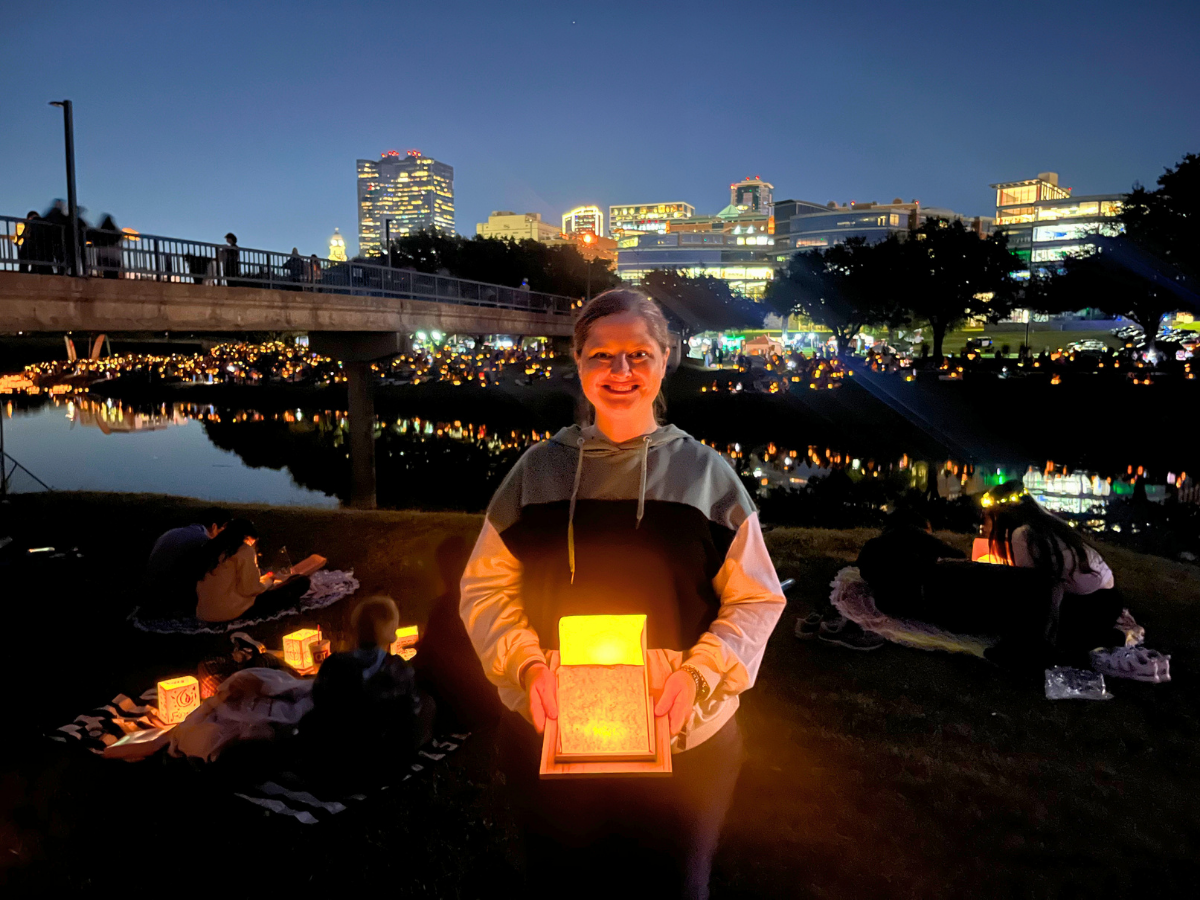 Woman in a gray hoodie orients a glowing lantern toward the camera at a nighttime riverside gathering, with city lights and a bridge in the background.