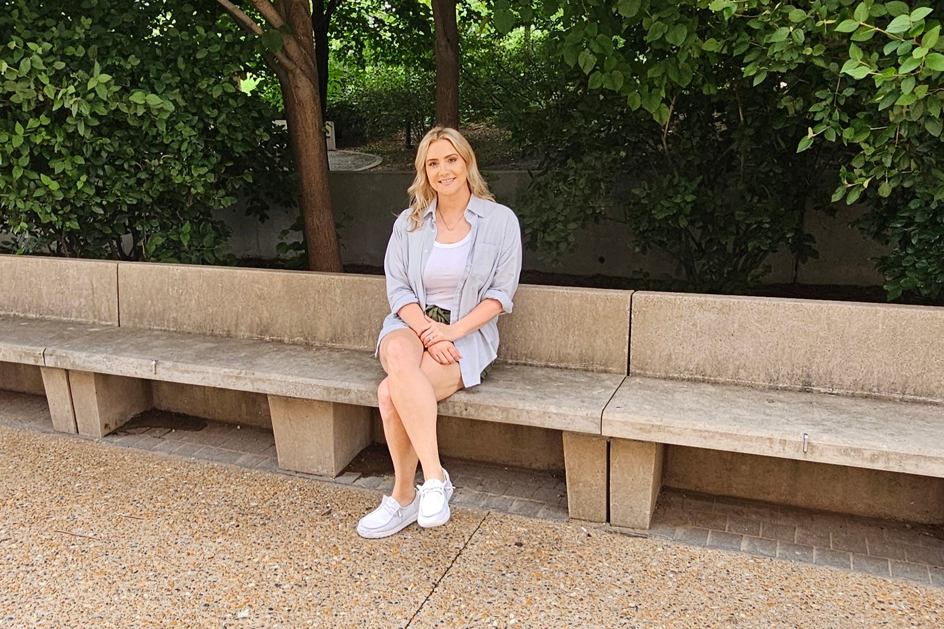 Woman sitting on a stone bench in a sunny park with green bushes behind her.