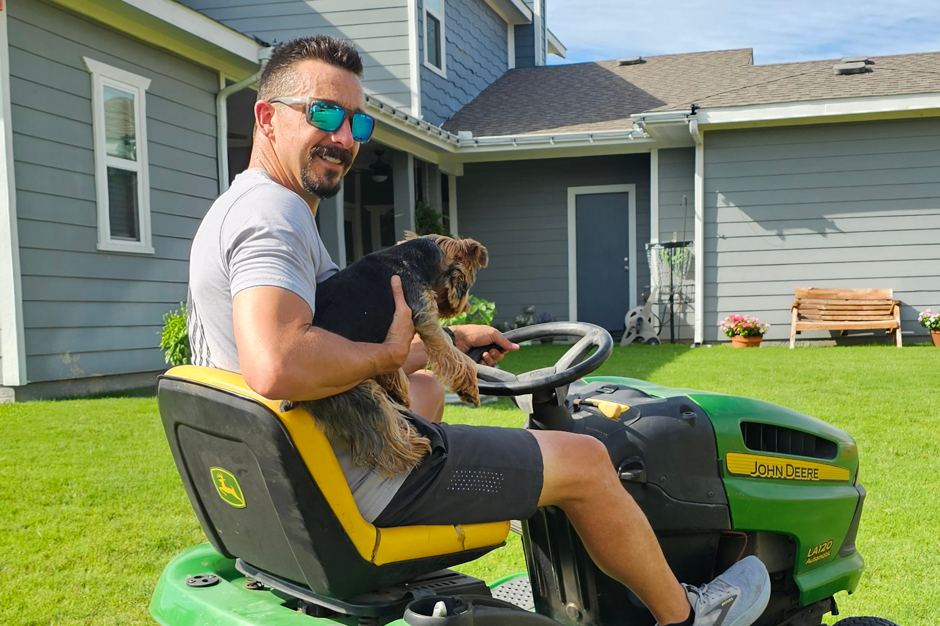 Man wearing sunglasses sits on a John Deere lawn tractor in a backyard, holding a small dog in his arms