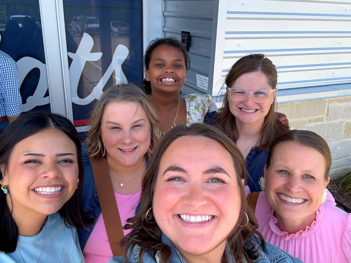 Six women of diverse backgrounds taking a group selfie, smiling at the camera outdoors near a building entrance.