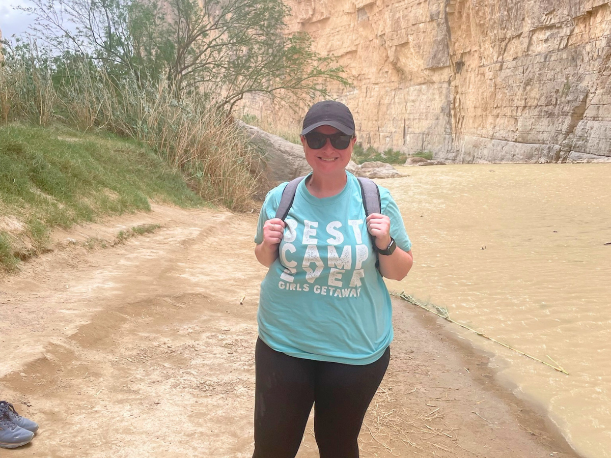 Smiling woman in a teal shirt with 'Best Camp Crew' text, wearing a cap and backpack, posing on a dirt trail beside a muddy river and rocky cliff wall.