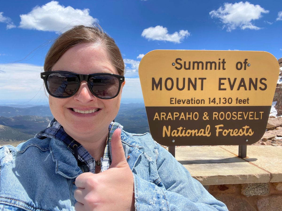 Smiling woman in sunglasses and a denim jacket gives a thumbs up beside the Mount Evans summit sign at 14,130 feet, with clear blue sky and distant mountains in the background.