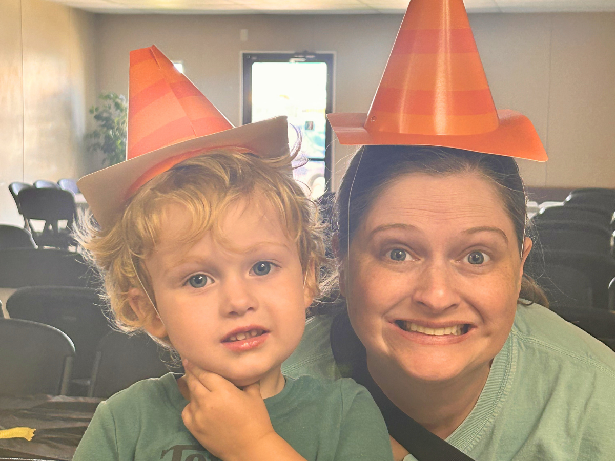 Woman and young boy wearing orange cone party hats, smiling at camera in a bright room with chairs in the background.