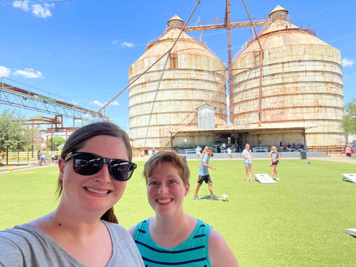 Two smiling women take a sunny selfie in front of huge rusted grain silos with a green lawn and people playing in the background.