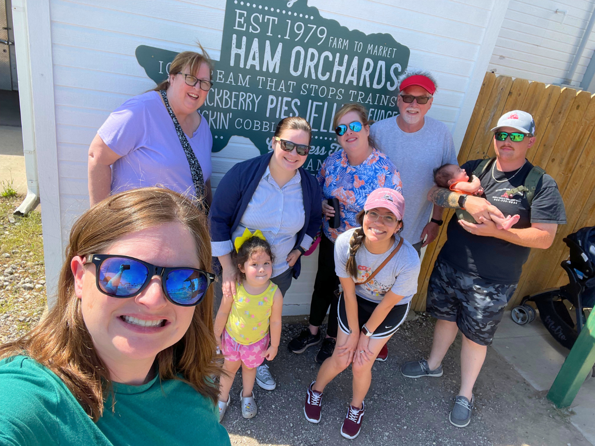 Group of eight people and two kids pose for a sunny outdoor selfie in front of a Ham Orchards sign (Est. 1979) on a blue wall, with a baby held by a man on the right.