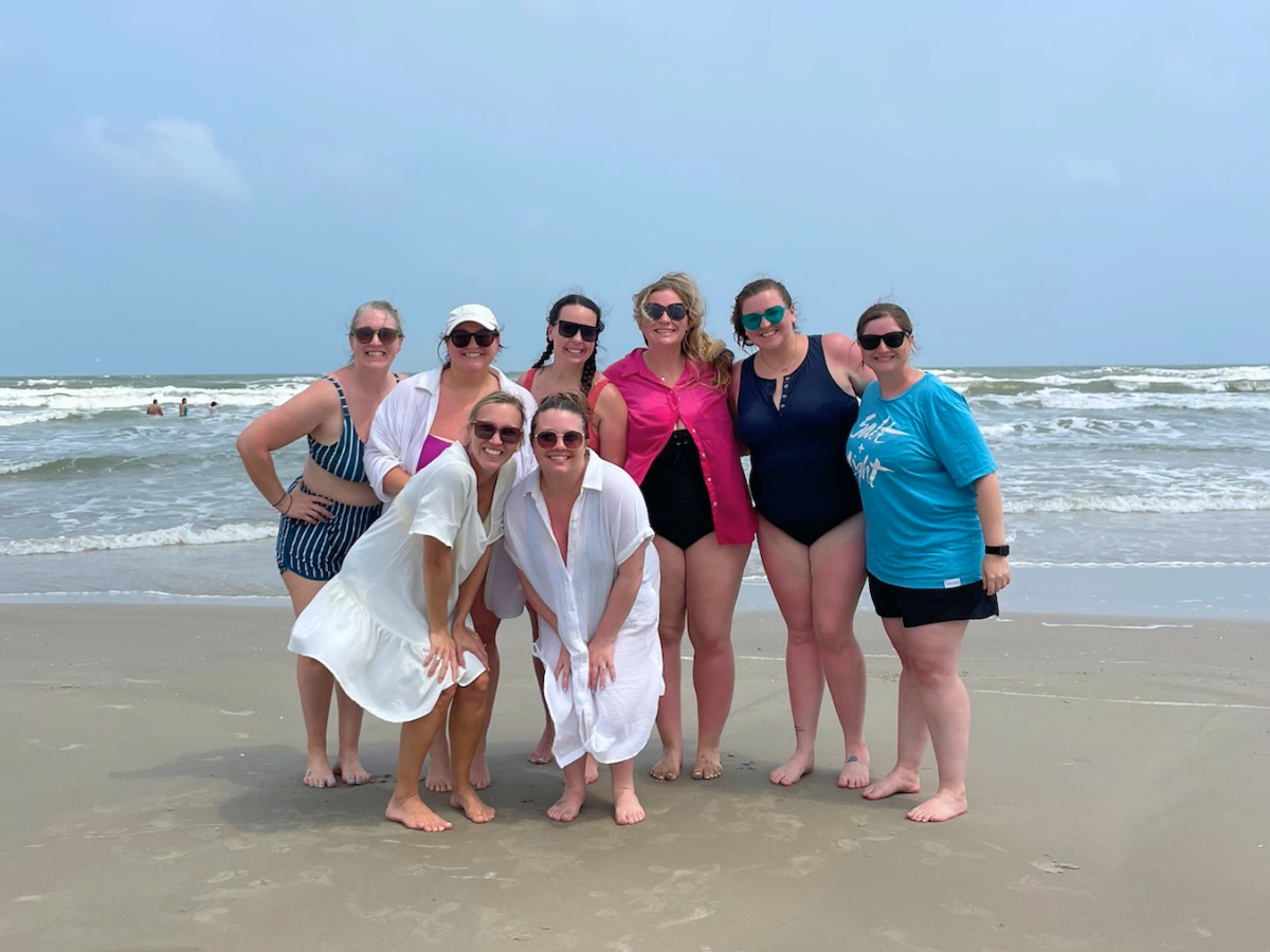 Eight women in swimsuits pose together on a sandy beach with waves behind them.