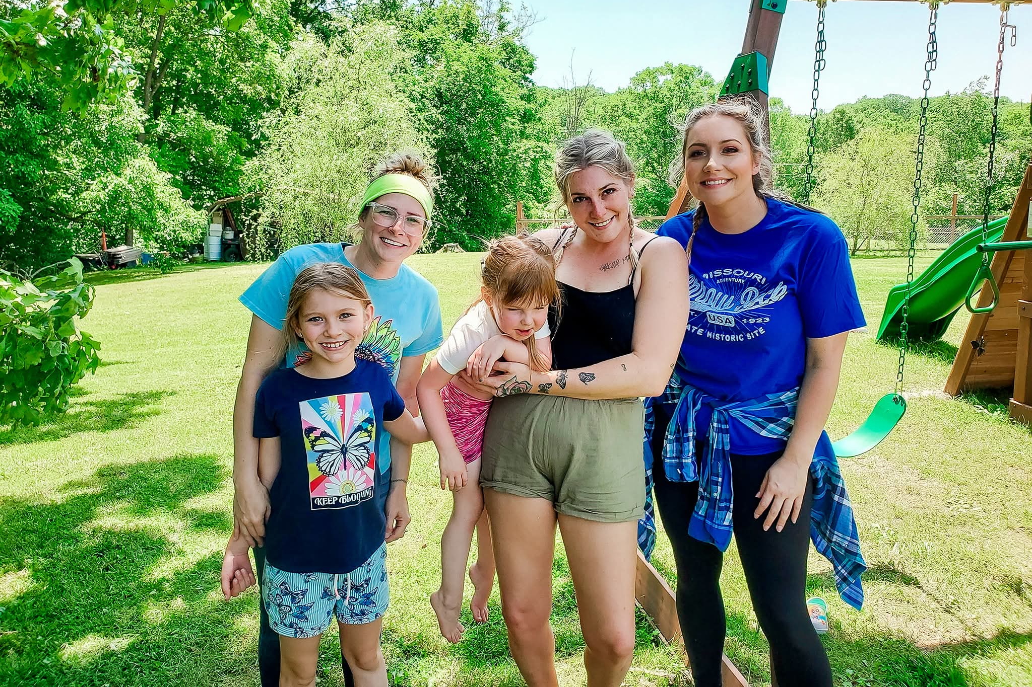 Five people smiling and posing for a photo in a sunny backyard with a wooden swing set and lush green trees nearby.