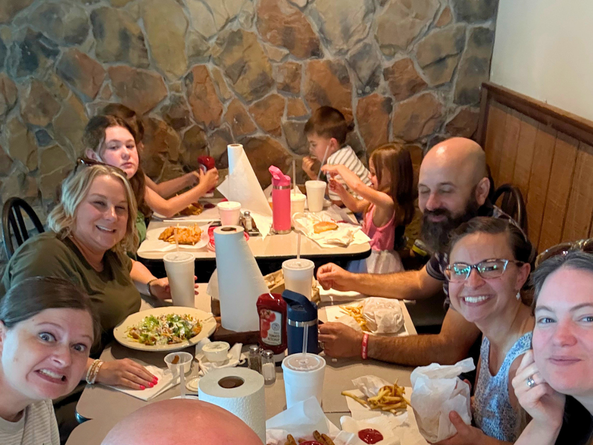 Group of nine people dining together at a casual restaurant, smiling at the camera with plates of fries and salads on the table.