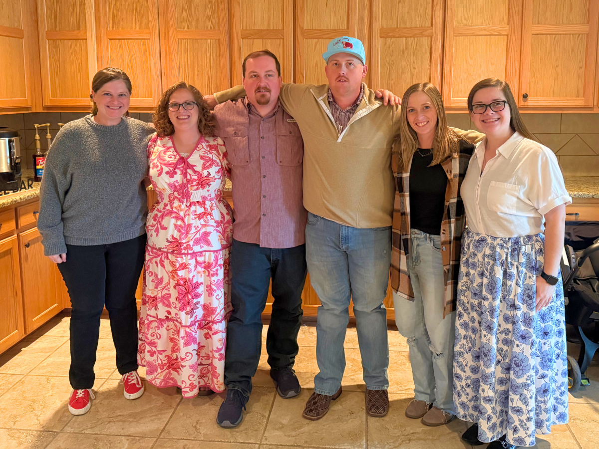 Six adults standing in a kitchen with wooden cabinets, smiling at the camera a group photo in a home setting (arms around each other).