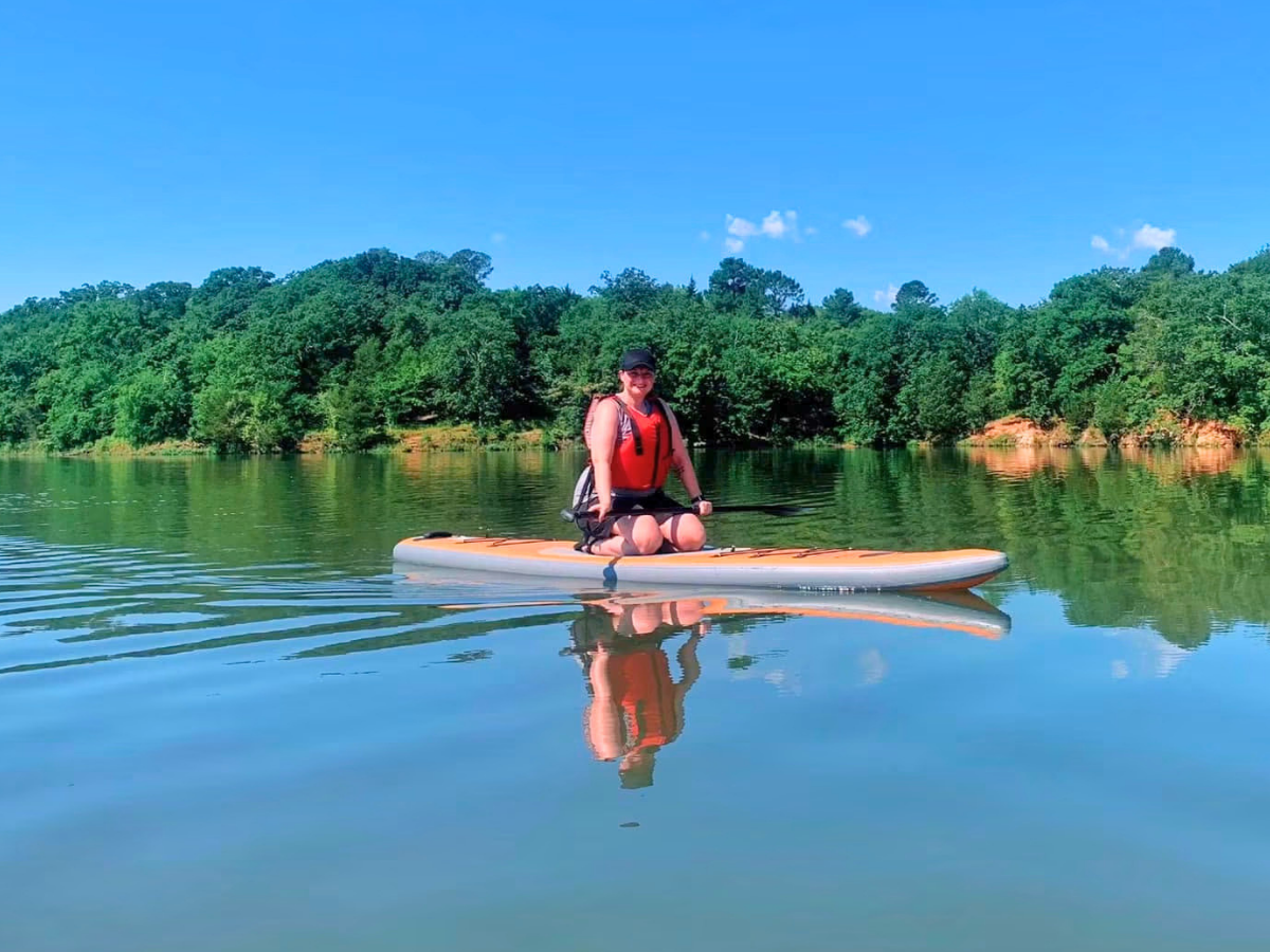 Person wearing a life jacket sits on an orange paddleboard on a calm lake with a tree-lined shore under a blue sky, reflection visible in the water
