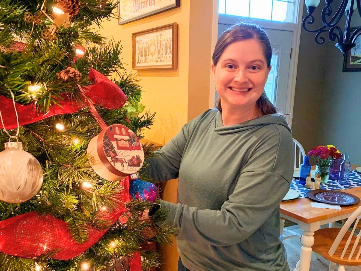Smiling woman decorates a Christmas tree with red ribbon and lights inside a cozy home.
