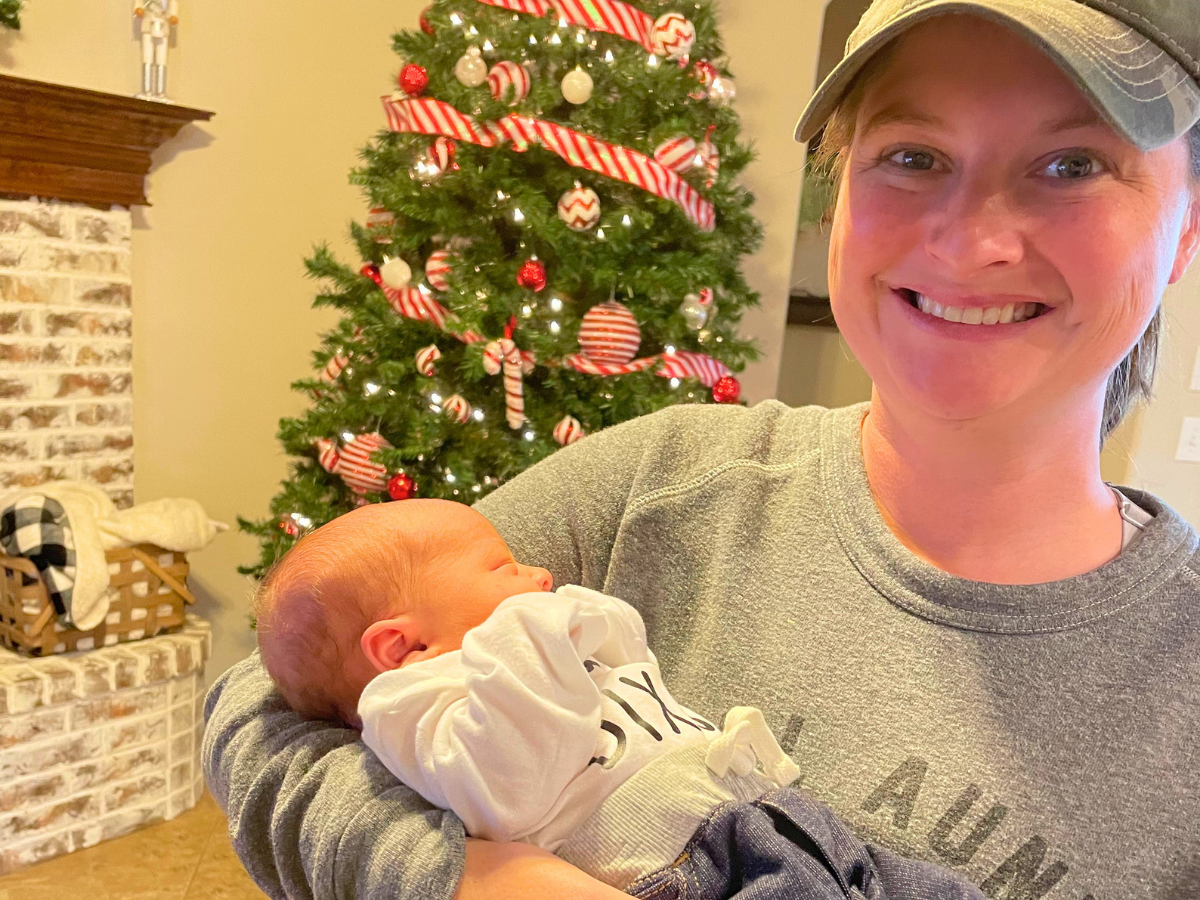Smiling woman in a cap holds a newborn baby in a cozy living room with a decorated Christmas tree in the background.