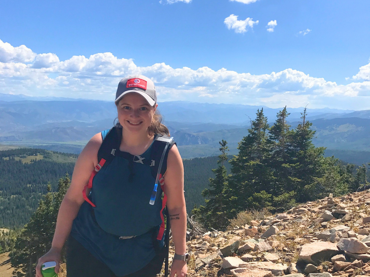 Woman hiker standing on a rocky mountain overlook with a teal backpack and a cap, smiling at the camera as distant mountains rise behind her.